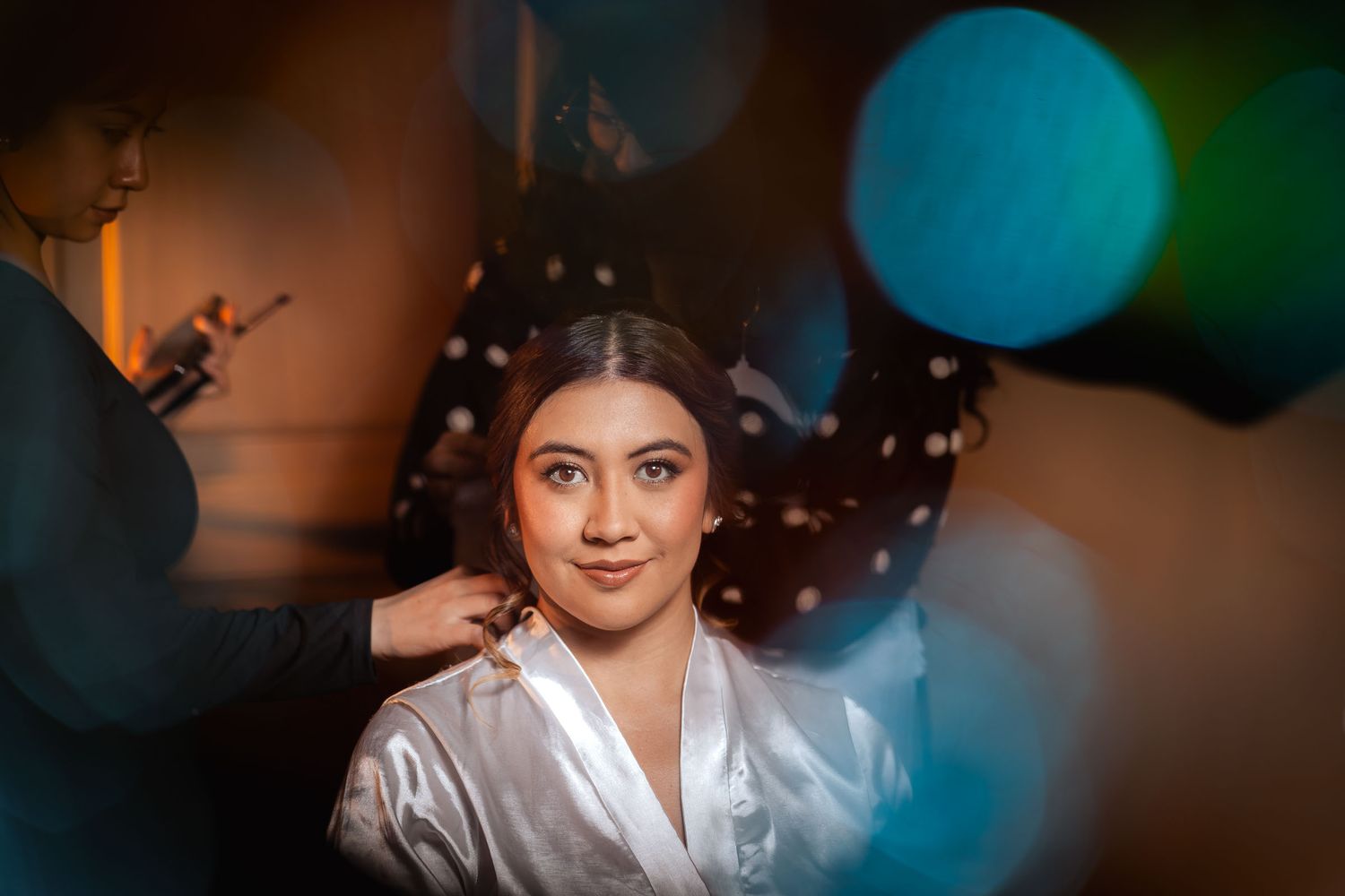 Novia en bata de satín mira a cámara mientras estilista arregla su peinado; preparación boda con luces bokeh.