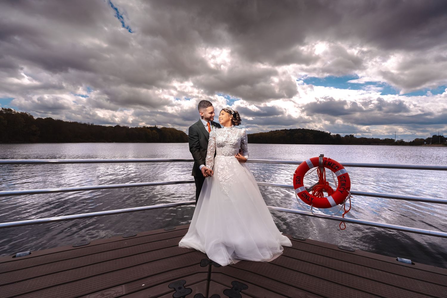 Romántica boda en Worcester: pareja abrazada en barco sobre lago con cielo nublado. Novia con vestido blanco, novio con traje oscuro