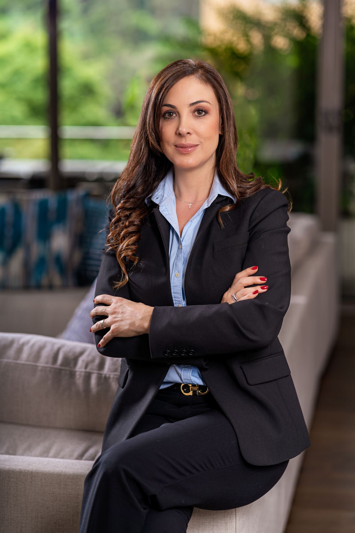 Mujer profesional con traje negro y camisa azul posa sentada con brazos cruzados; retrato corporativo. Sandra Vélez