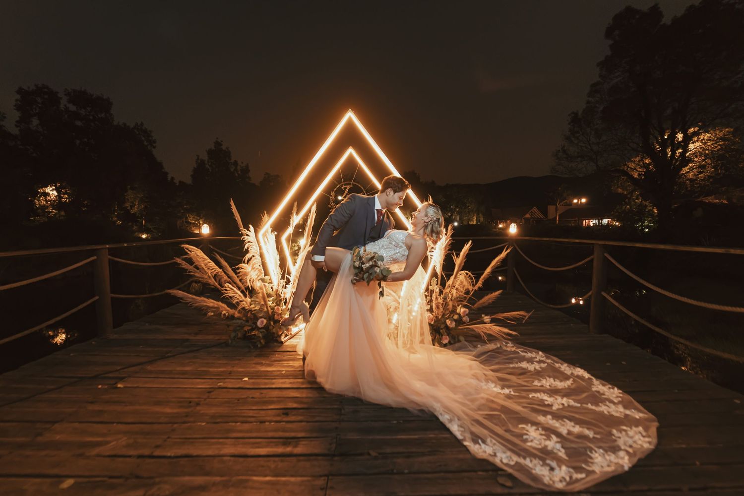 Boda nocturna romántica: pareja besándose bajo arco de neón y decoración boho en puente de madera. Ambiente cálido y mágico en la oscuridad
