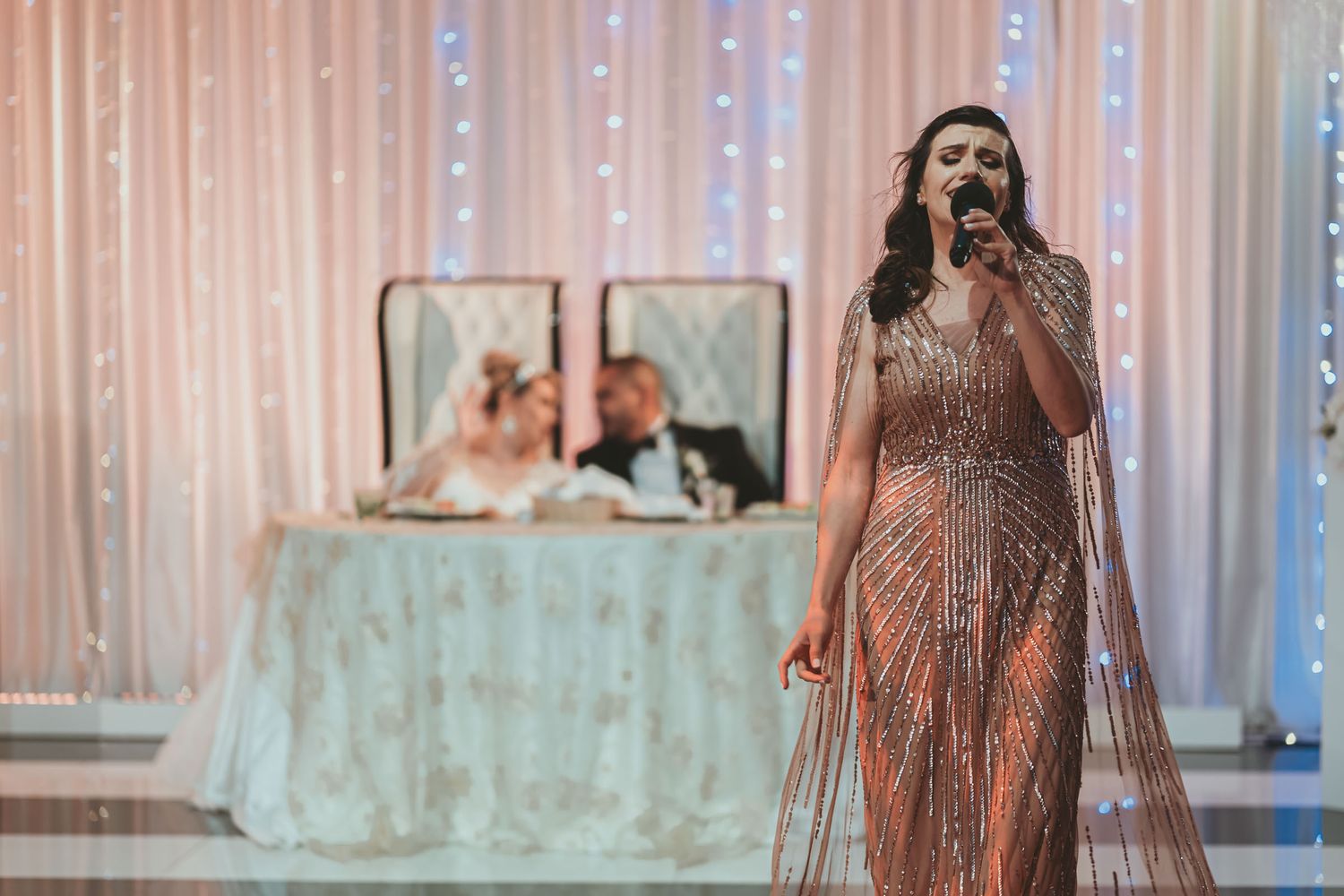 Cantante con vestido brillante actúa para novios sentados en mesa principal; recepción boda, cortina luces.
