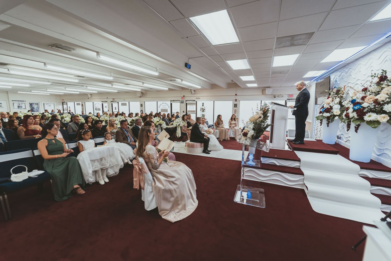 Ceremonia boda en progreso; oficiante habla desde púlpito a novios e invitados en salón amplio.