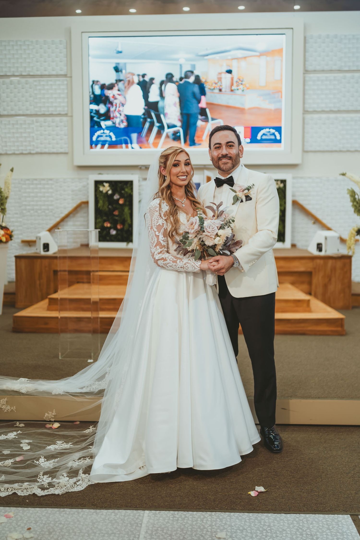 Novios posan abrazados en altar de iglesia decorado; ella sostiene ramo. Retrato boda formal.