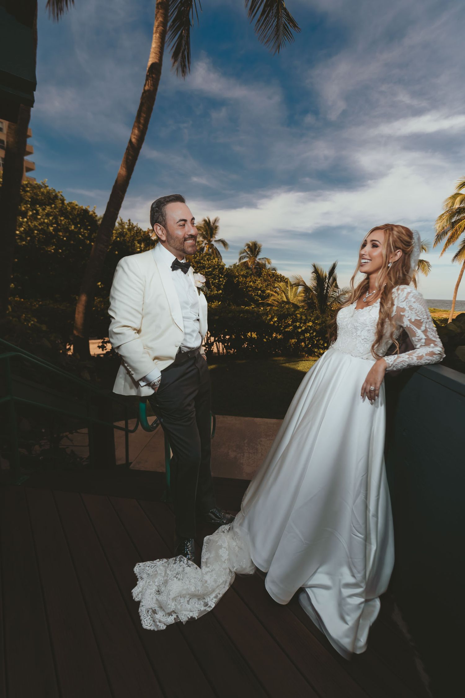 Novios posan elegantes en balcón con palmeras y cielo azul; él chaqueta clara, ella vestido blanco.