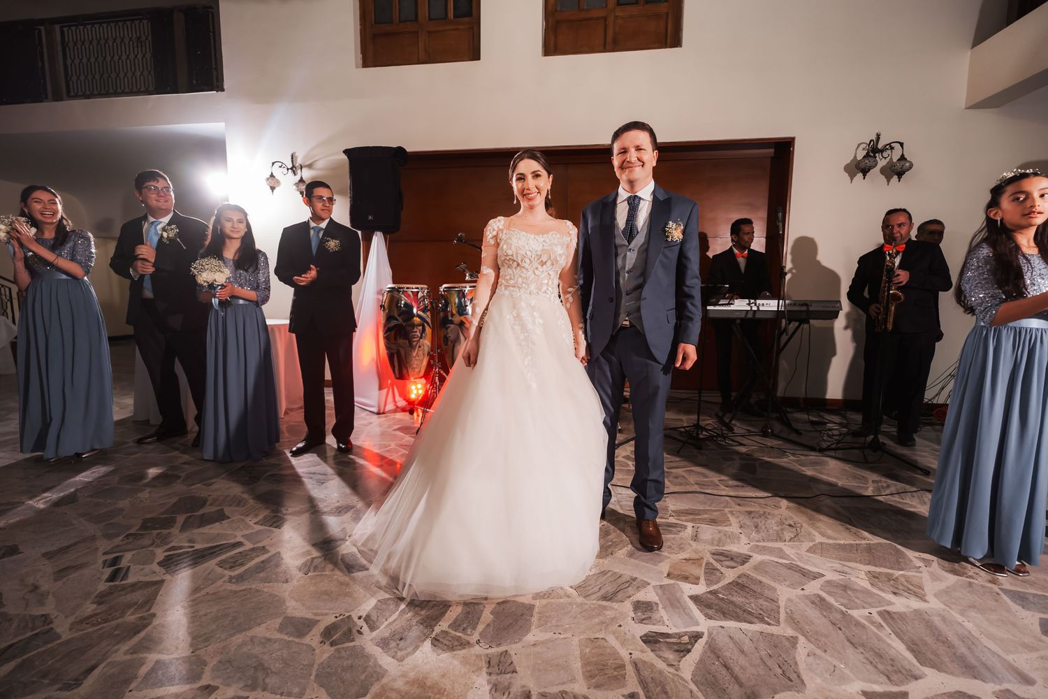 Novios sonrientes posan en centro de salón de recepción flanqueados por cortejo con velas encendidas; boda.