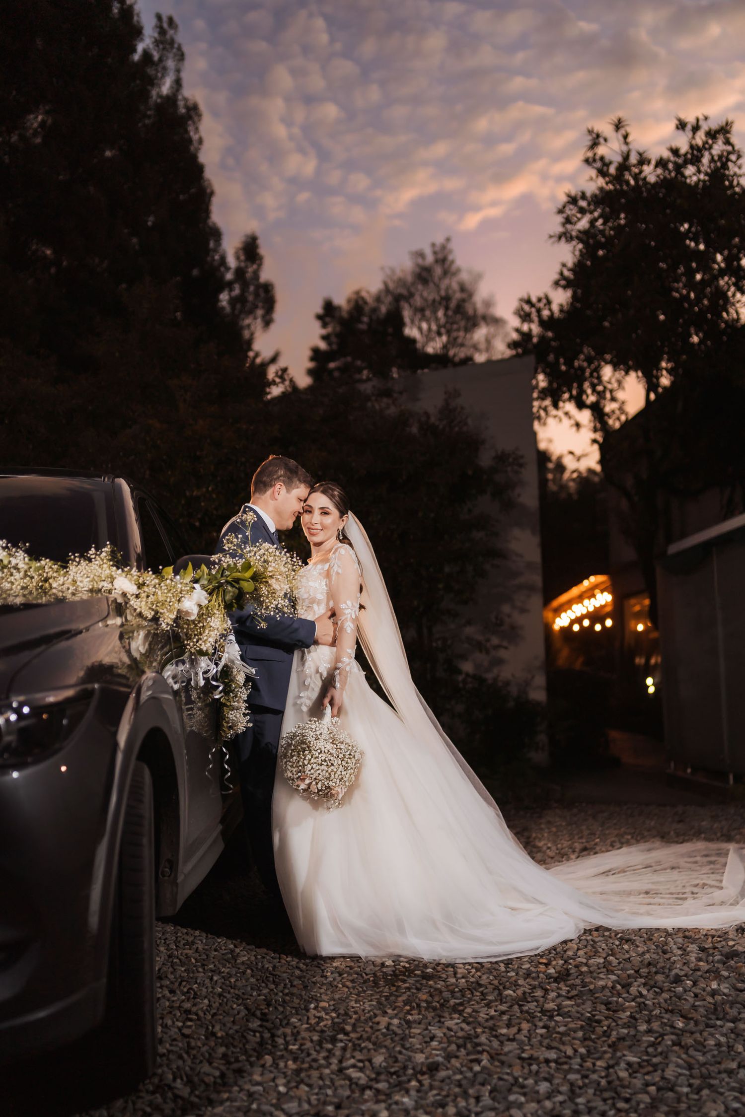 Pareja de novios posa junto a coche oscuro decorado para boda al atardecer; novia sostiene ramo, velo largo.