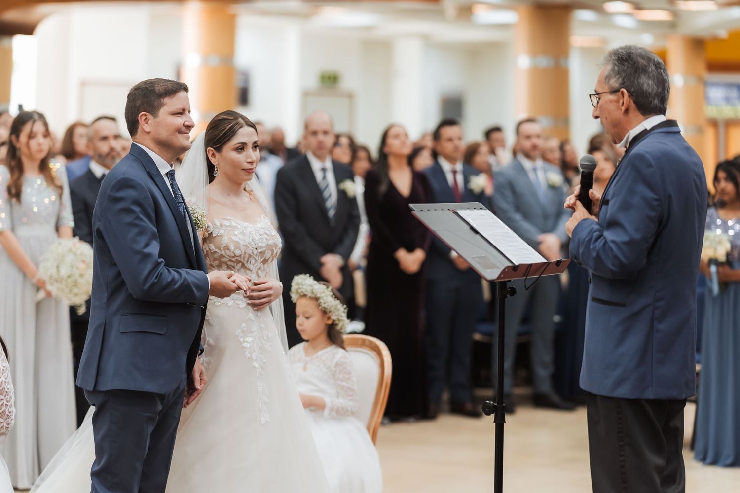 Novios escuchan atentos al oficiante durante ceremonia de boda IDMJI; invitados observan desde asientos.