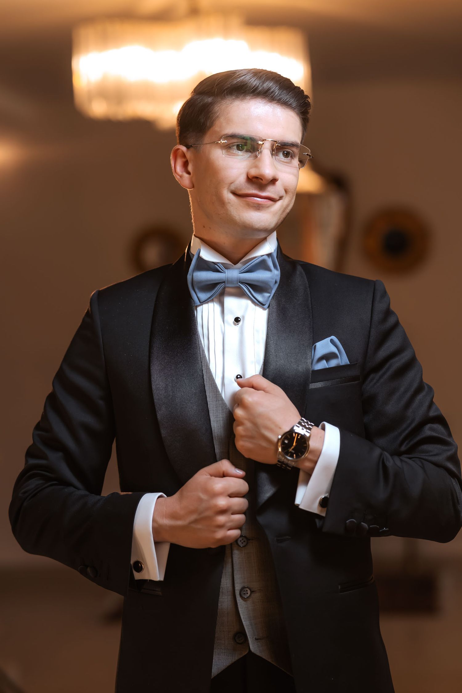 Retrato del novio ajustando su esmoquin negro con chaleco gris y pajarita azul; preparación de boda en interior elegante.
