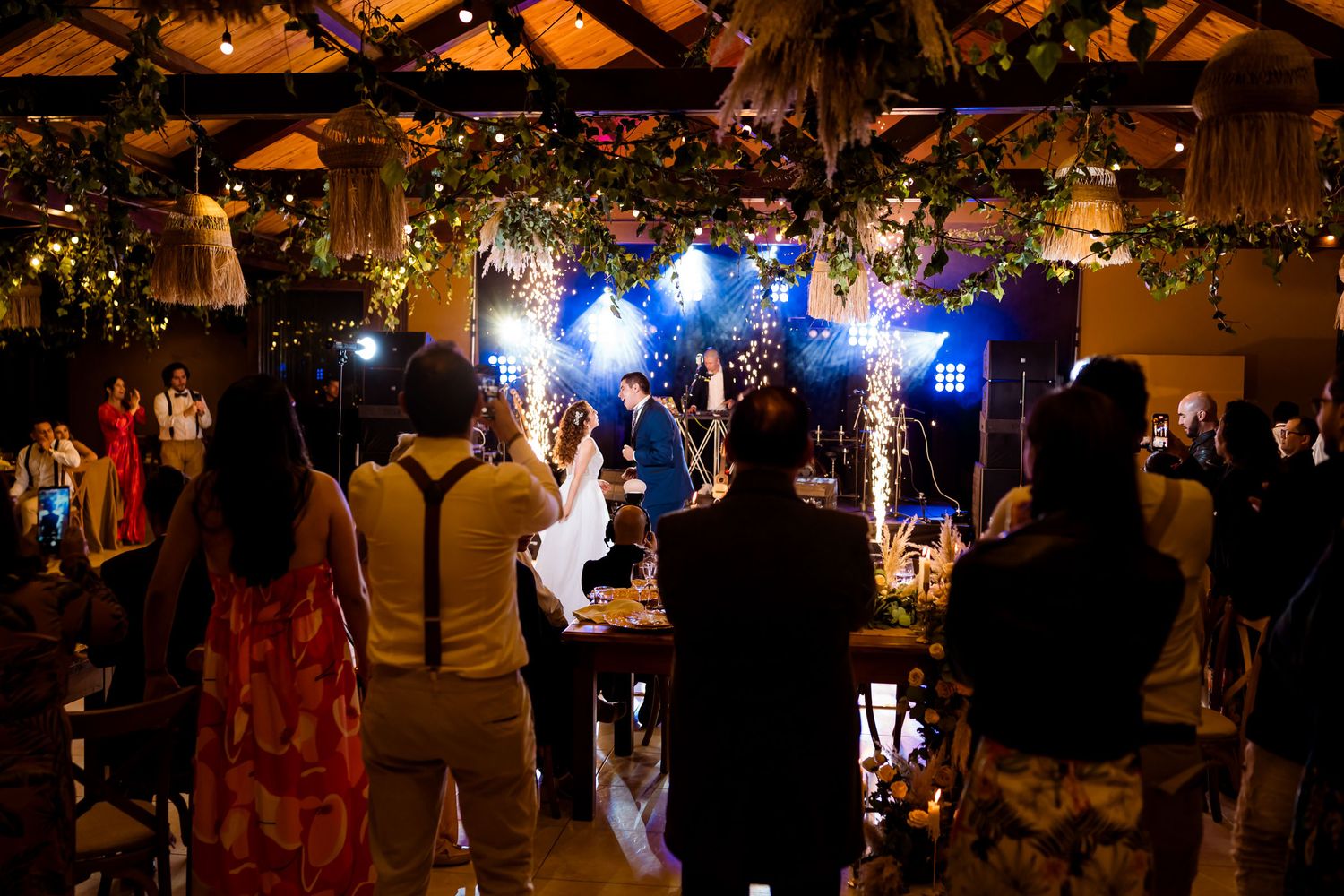 Invitados observan y fotografían a novios bailando en escenario con pirotecnia fría; recepción boda rústica.