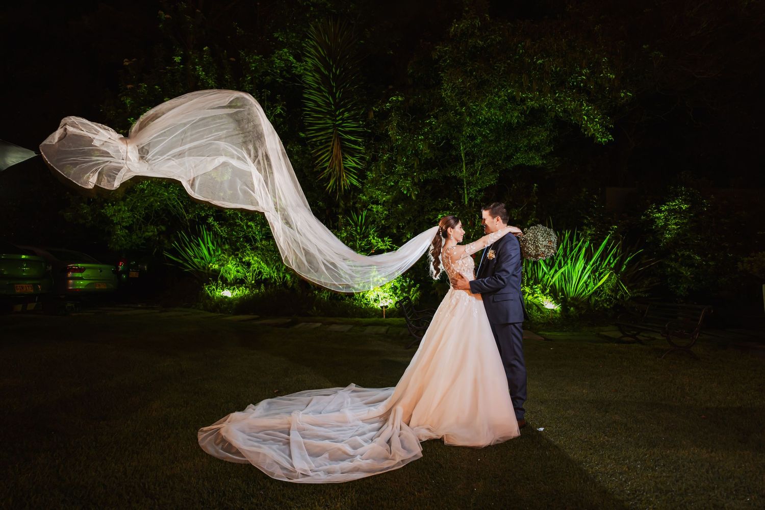 Novios posan de noche en jardín iluminado; velo largo de novia flota espectacularmente. Boda mágica.