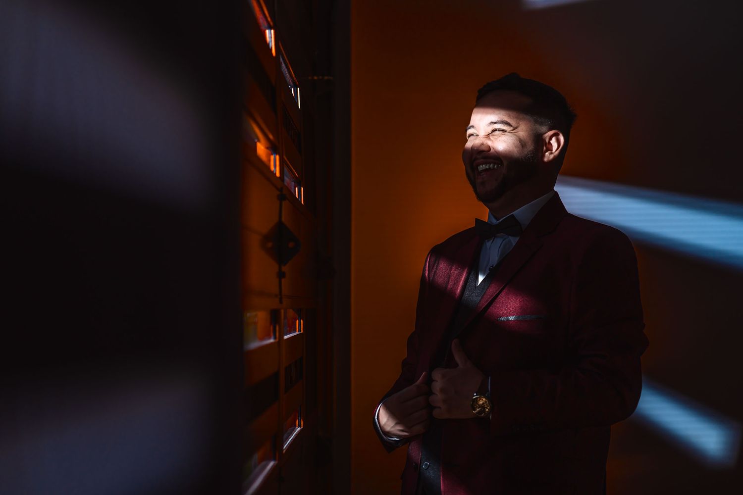 Retrato novio sonriente con traje vino tinto en habitación oscura; haces de luz diagonal crean sombras. Boda.