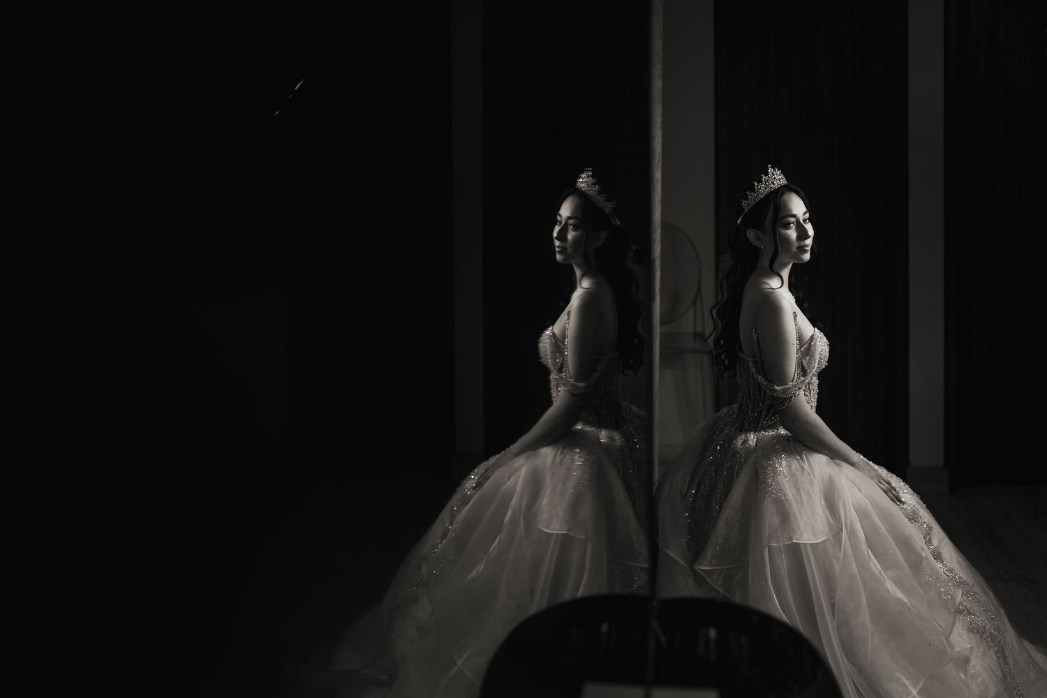 Quinceañera sentada de perfil con vestido y tiara, reflejada en espejo; fotografía artística en blanco y negro