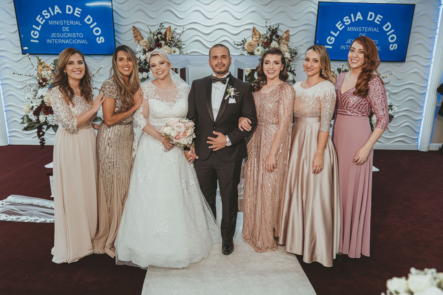 Novios posan con cortejo femenino (damas honor) en iglesia (IDMJI); vestidos largos elegantes.