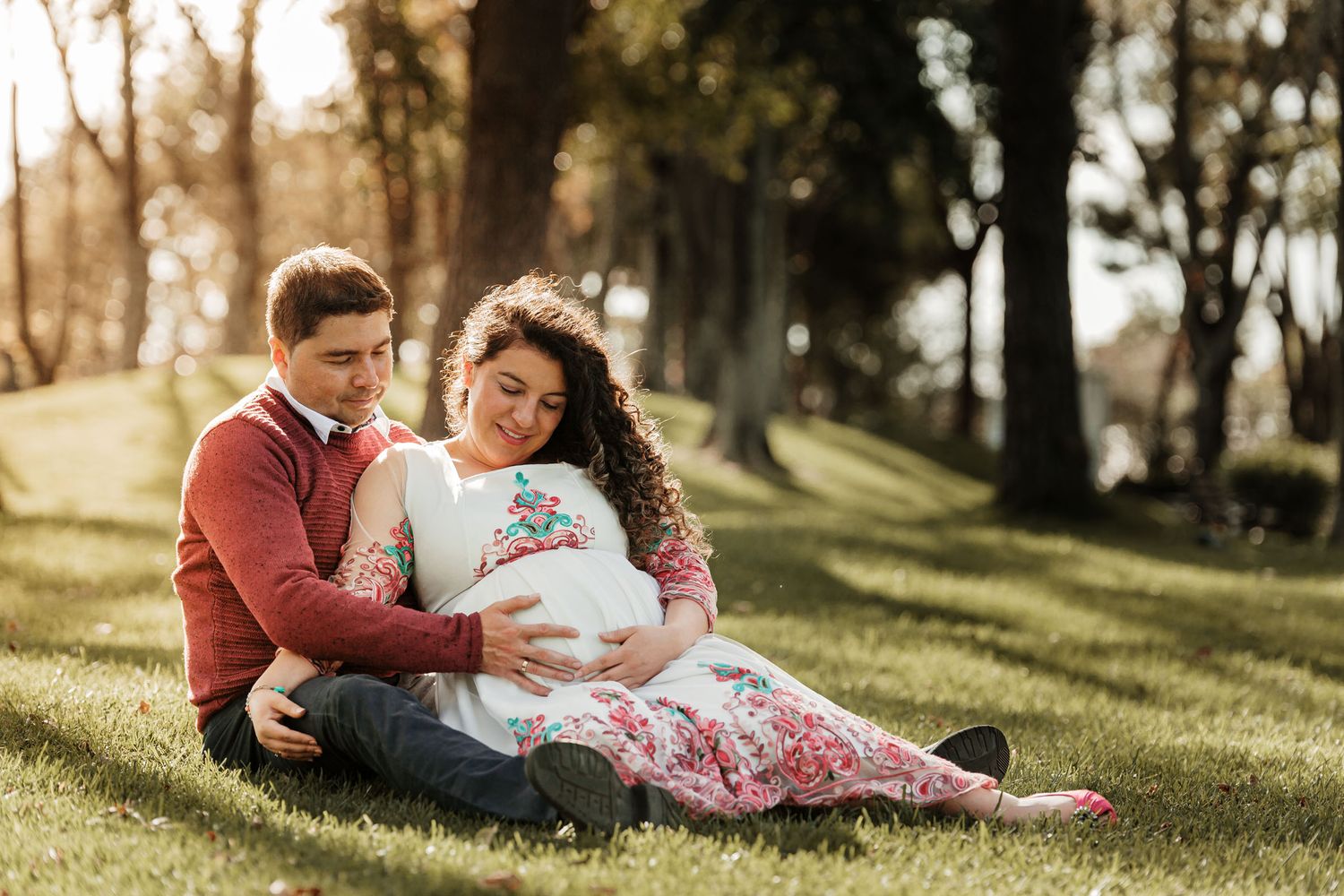 Pareja sentada en el césped abraza barriga de embarazo al atardecer. Foto de maternidad en parque.