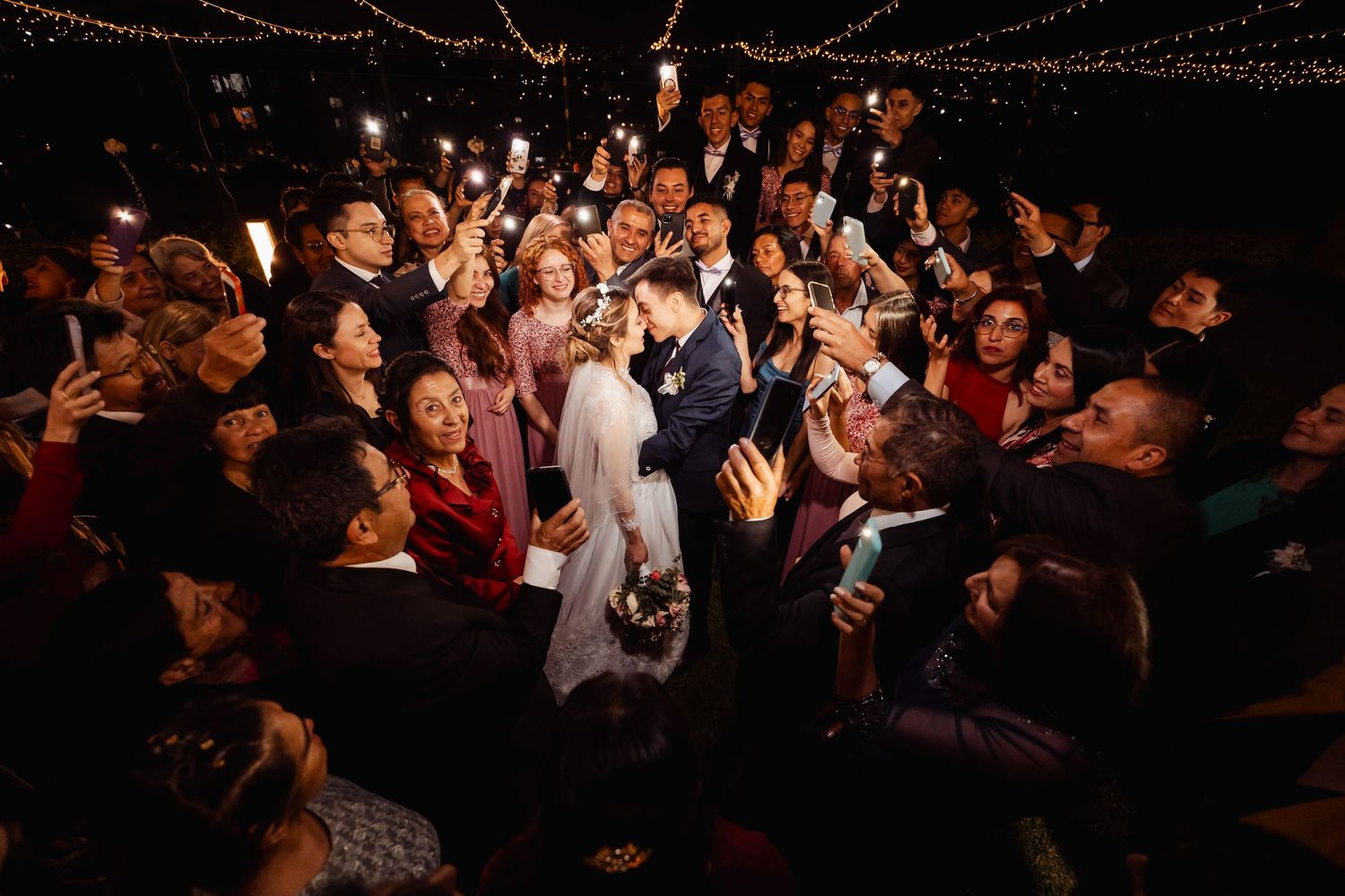 Novios se besan rodeados de invitados con móviles y luces encendidas; fiesta boda nocturna, vista superior.
