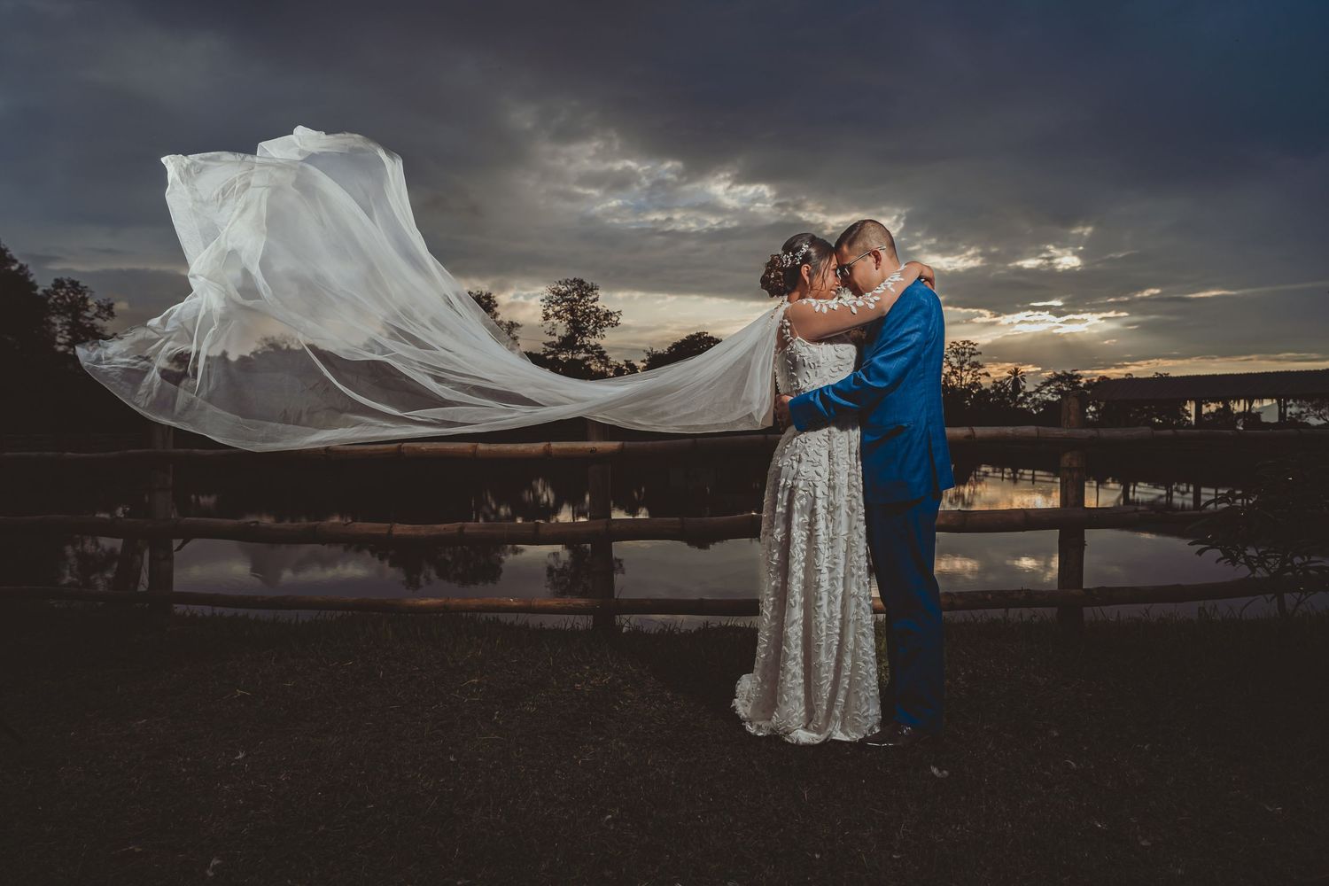 Novios se abrazan al atardecer junto a lago; velo largo de novia ondea espectacularmente al viento. Boda.