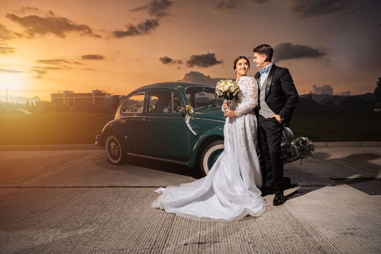 Novios sonríen posando junto a coche verde clásico en la puesta de sol. Foto de boda romántica exterior.
