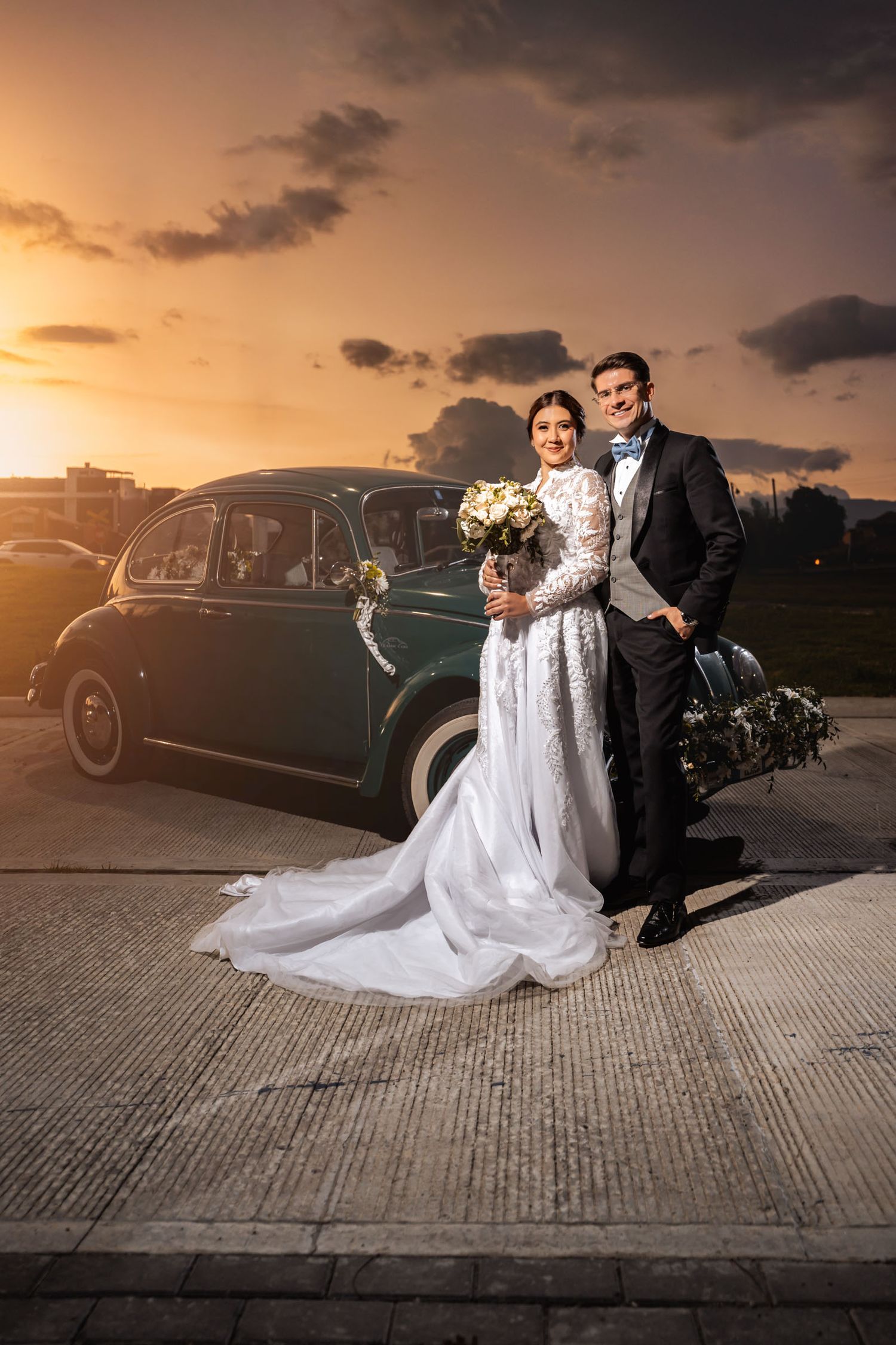 Novios sonríen posando junto a coche verde clásico en la puesta de sol. Foto de boda romántica exterior.