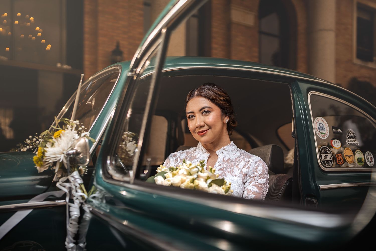 Novia con vestido de encaje y ramo mira a cámara desde interior de coche clásico VW Escarabajo verde; puerta abierta. Boda.