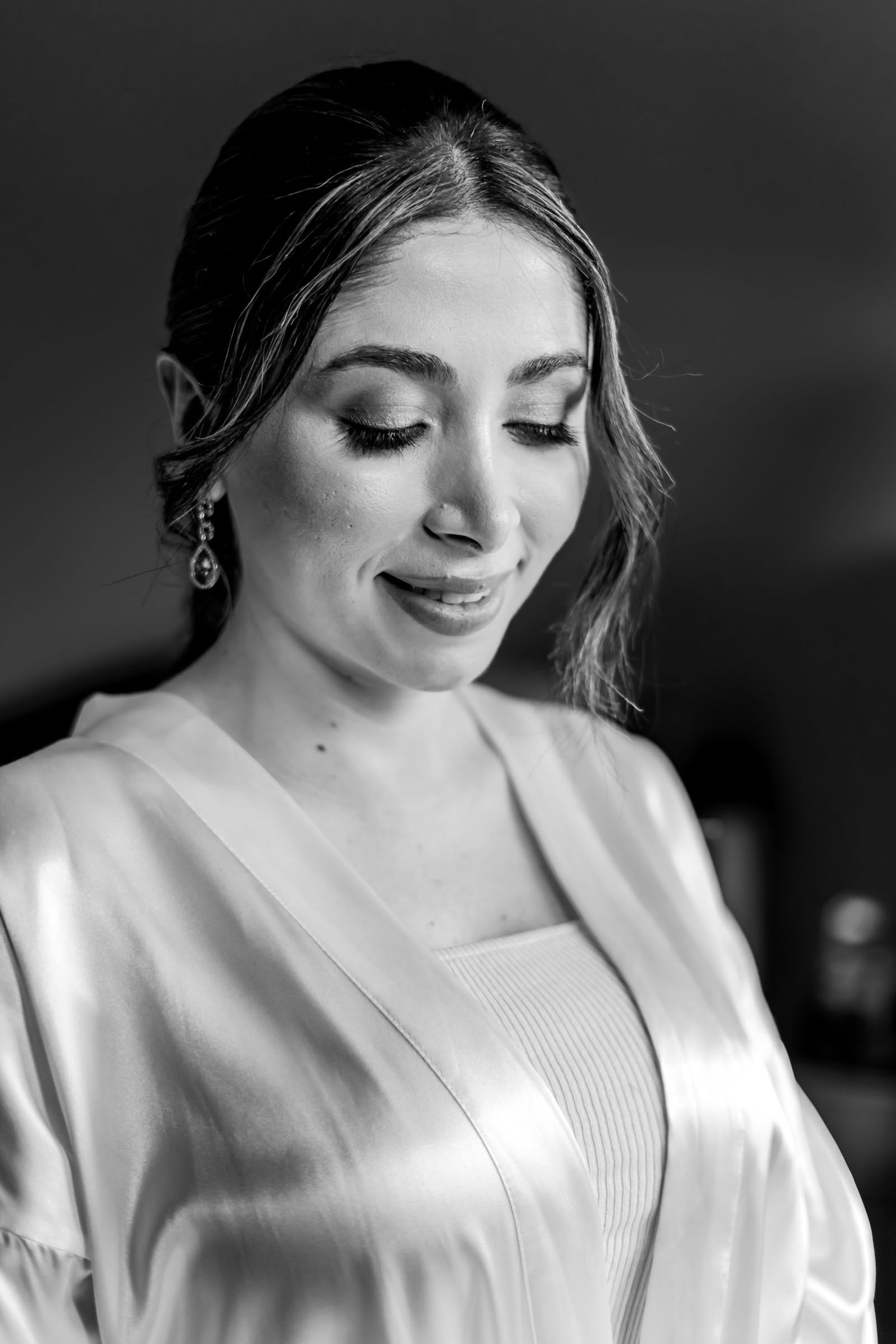 Retrato B&W novia con bata; mira hacia abajo sonriendo suavemente. Maquillaje elegante; preparación boda.