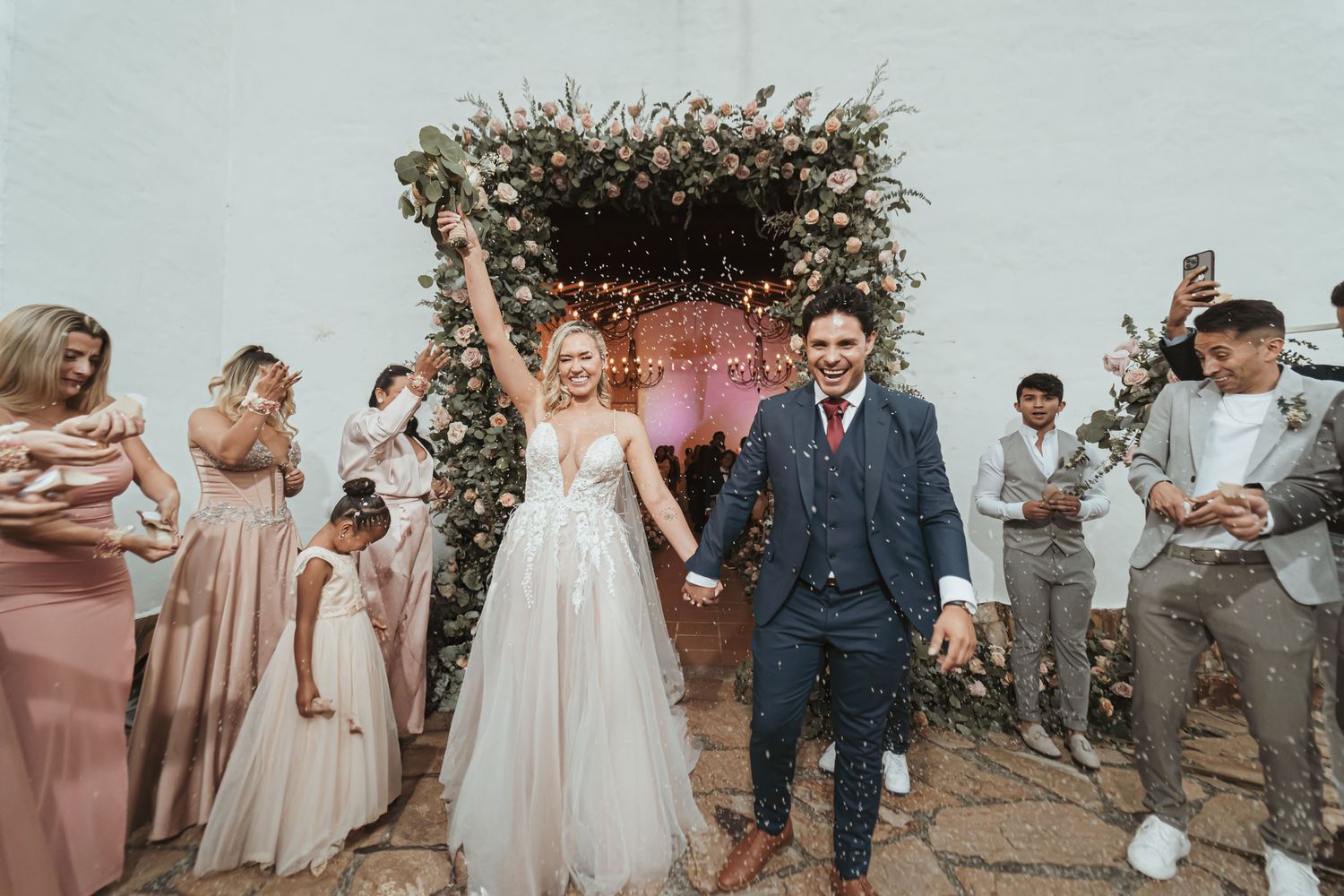 Novios celebran bajo lluvia de confeti al salir por arco floral; novia alza ramo, invitados alegres. Boda.