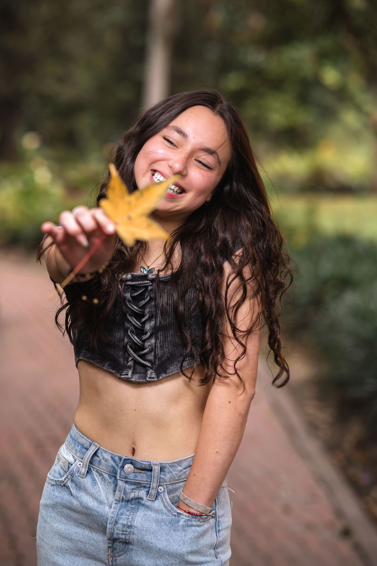 Joven sonriente con top negro y jeans muestra hoja otoñal amarilla en camino de parque; sesión prequince.