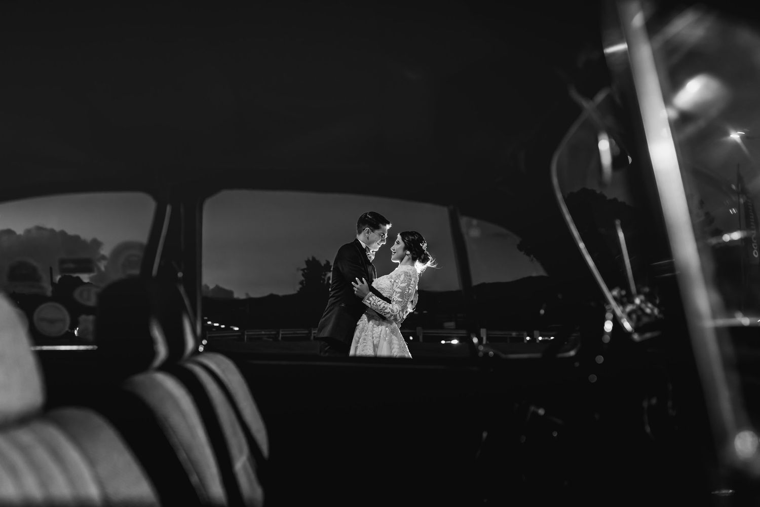 Beso romántico de novios en la oscuridad, vistos desde interior de coche. Fotografía de boda blanco y negro íntima.