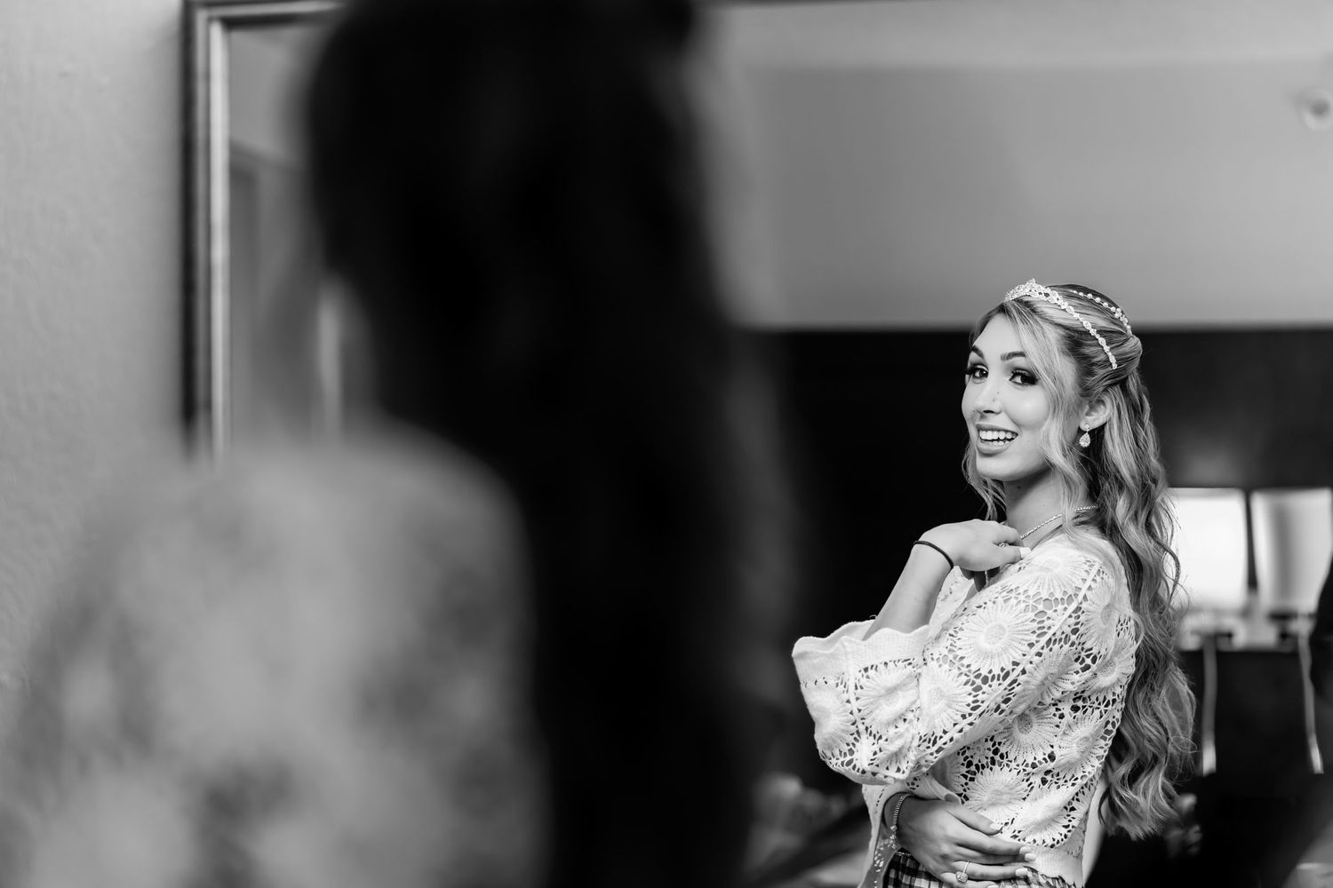 Foto B&W: Novia sonriente con bata de encaje y tocado mira su reflejo en espejo; preparación boda.