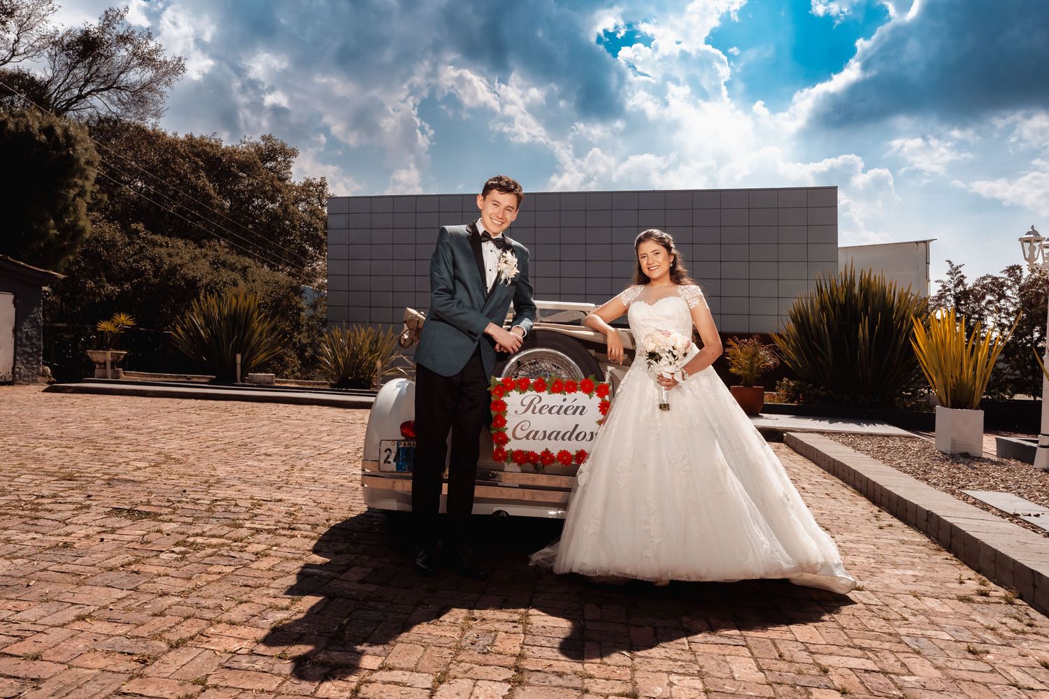 Novios sonrientes junto a coche de boda clásico en exterior moderno, cielo azul de fondo