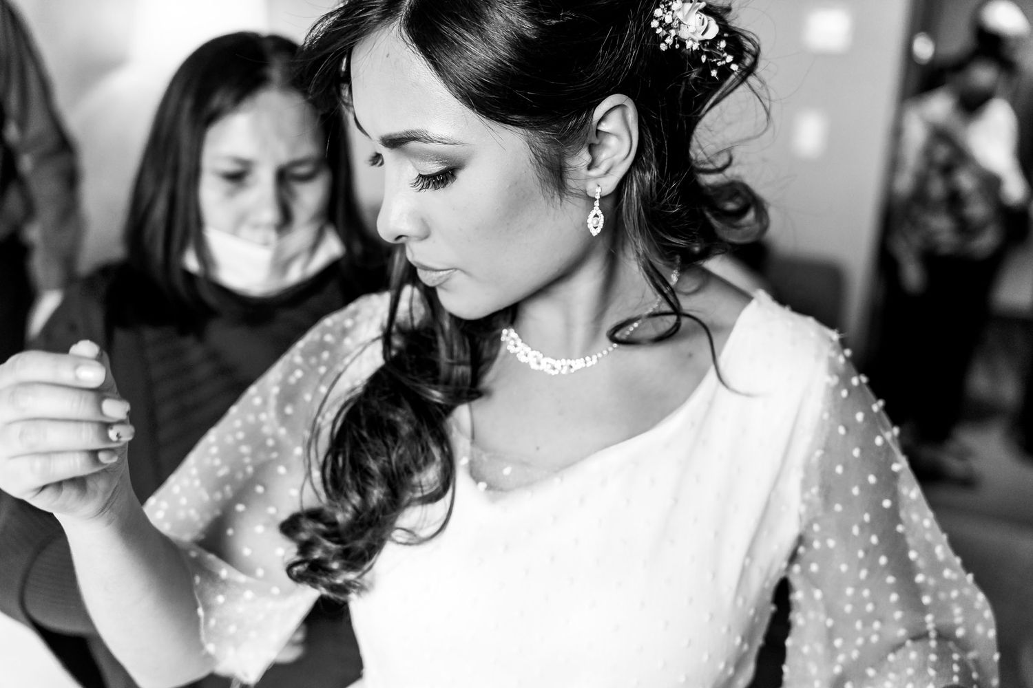Foto B&W: Novia baila con vestido manga larga (perlas?) durante recepción de boda; invitada fondo.