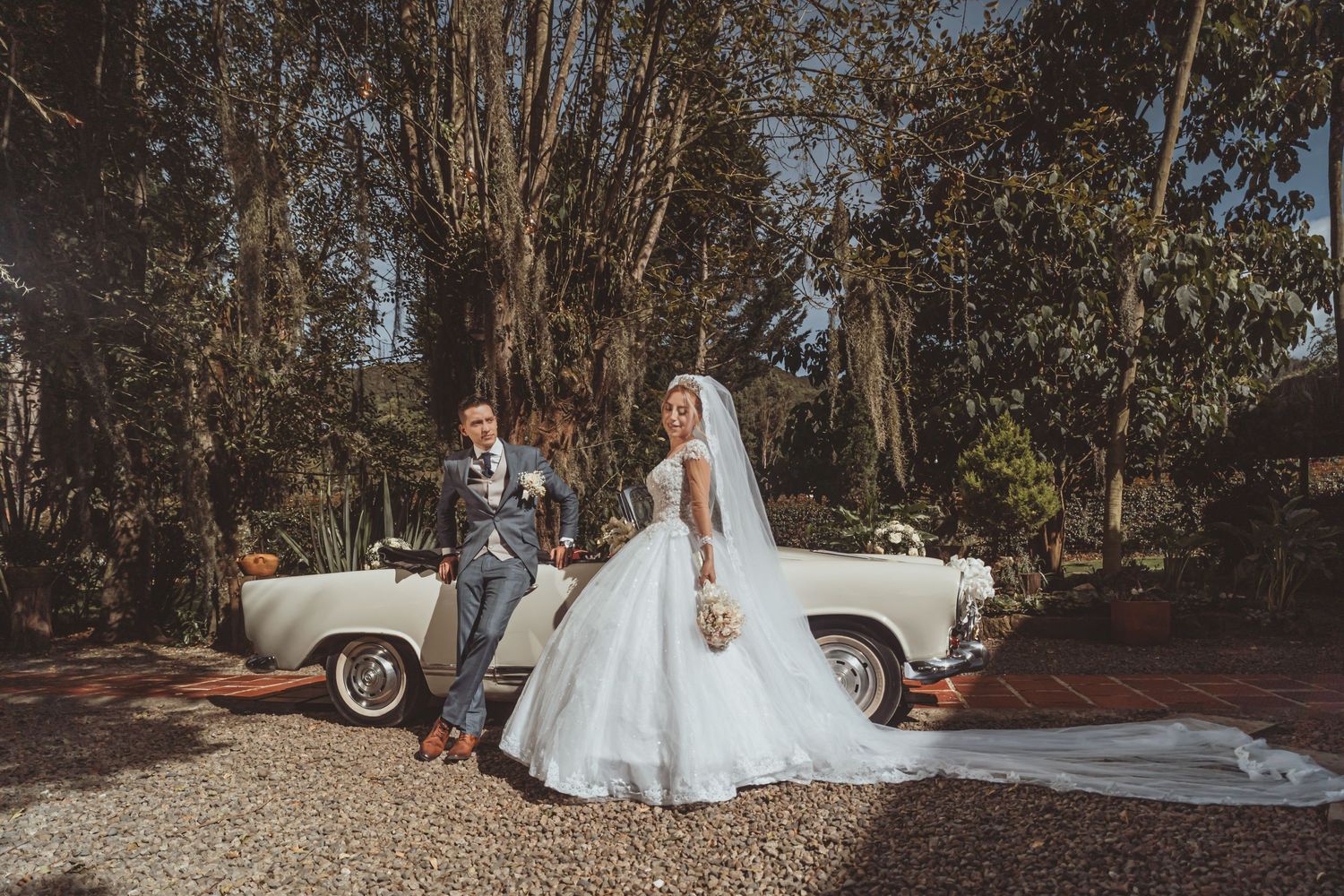 Novios posan elegantes junto a coche clásico convertible blanco en jardín soleado; novia velo largo. Boda.