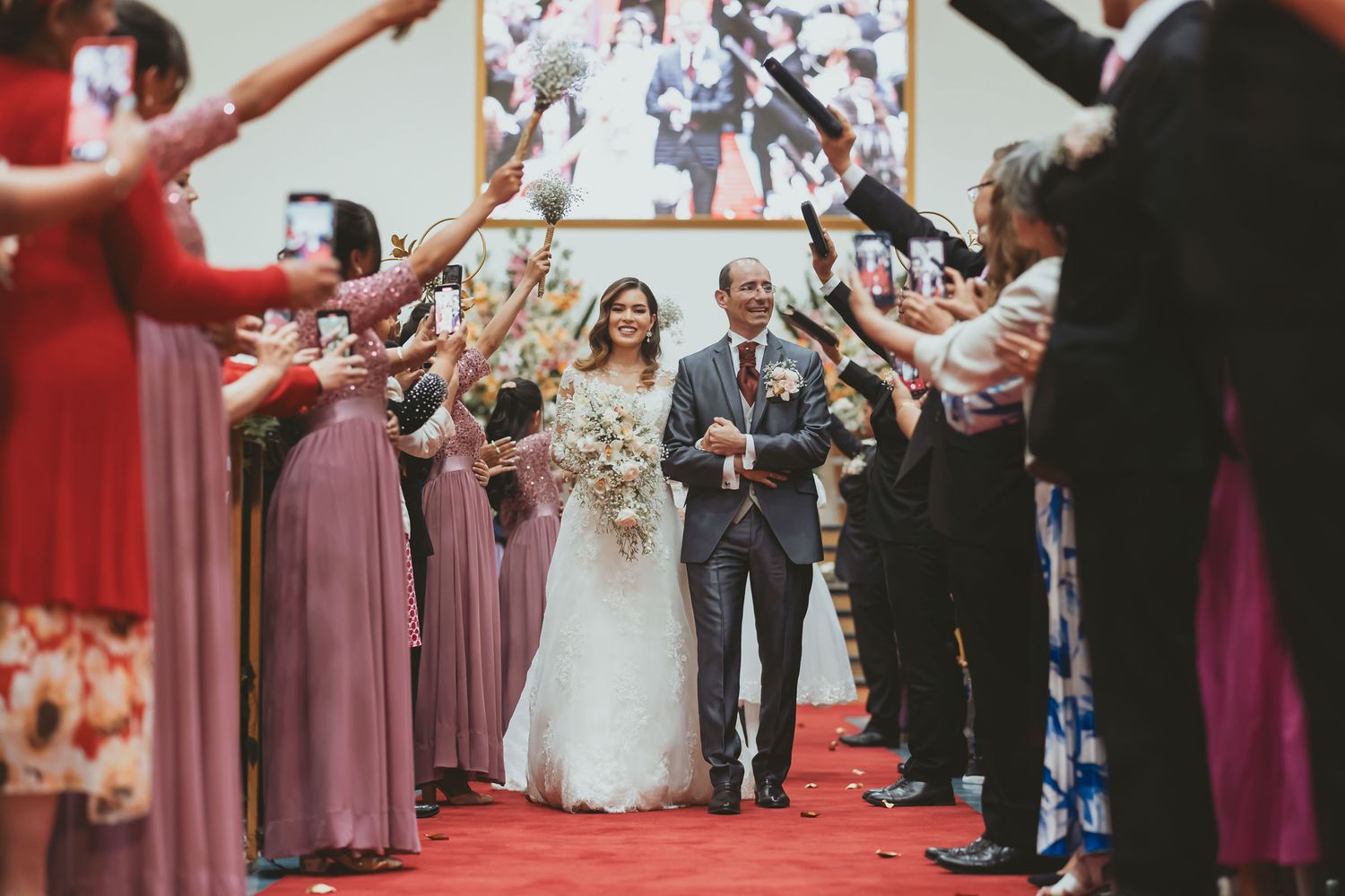 Novios caminan sonrientes por pasillo de alfombra roja, invitados celebran su paso levantando flores. Boda IDMJI
