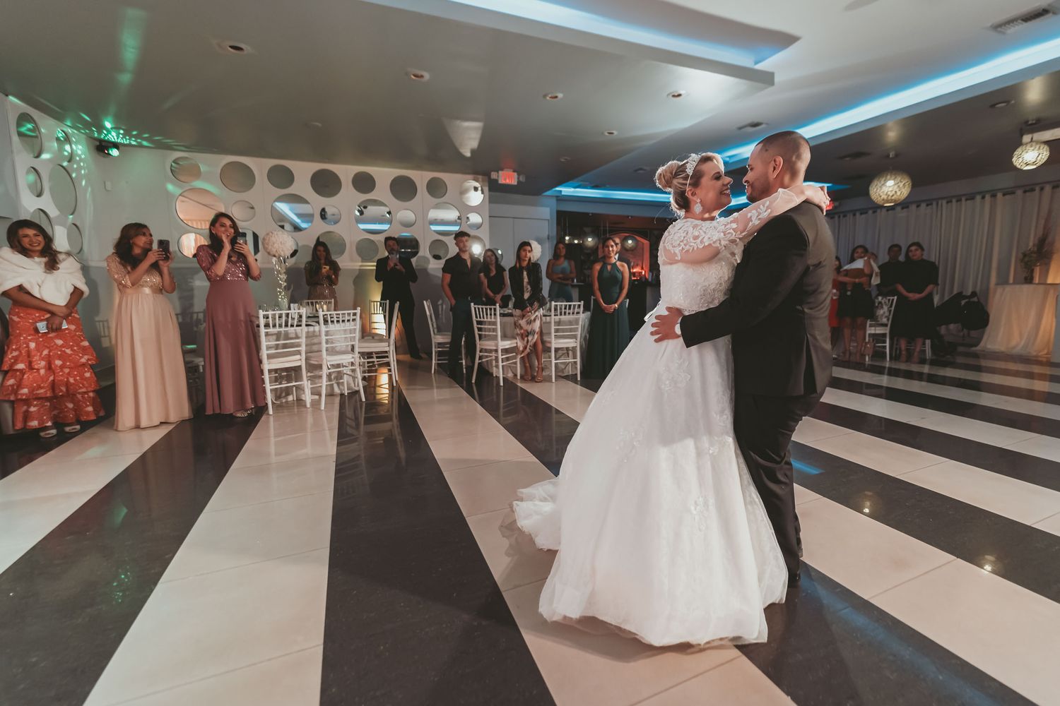 Novios comparten primer baile romántico en pista a cuadros; invitados aplauden al fondo. Recepción boda.