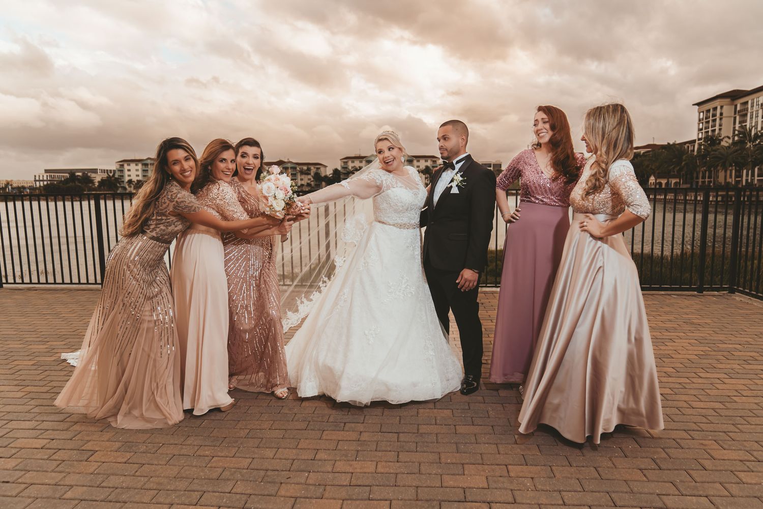 Novios posan con sus damas de honor (vestidos rosa/champagne) en terraza al atardecer; foto grupal boda en Miami.