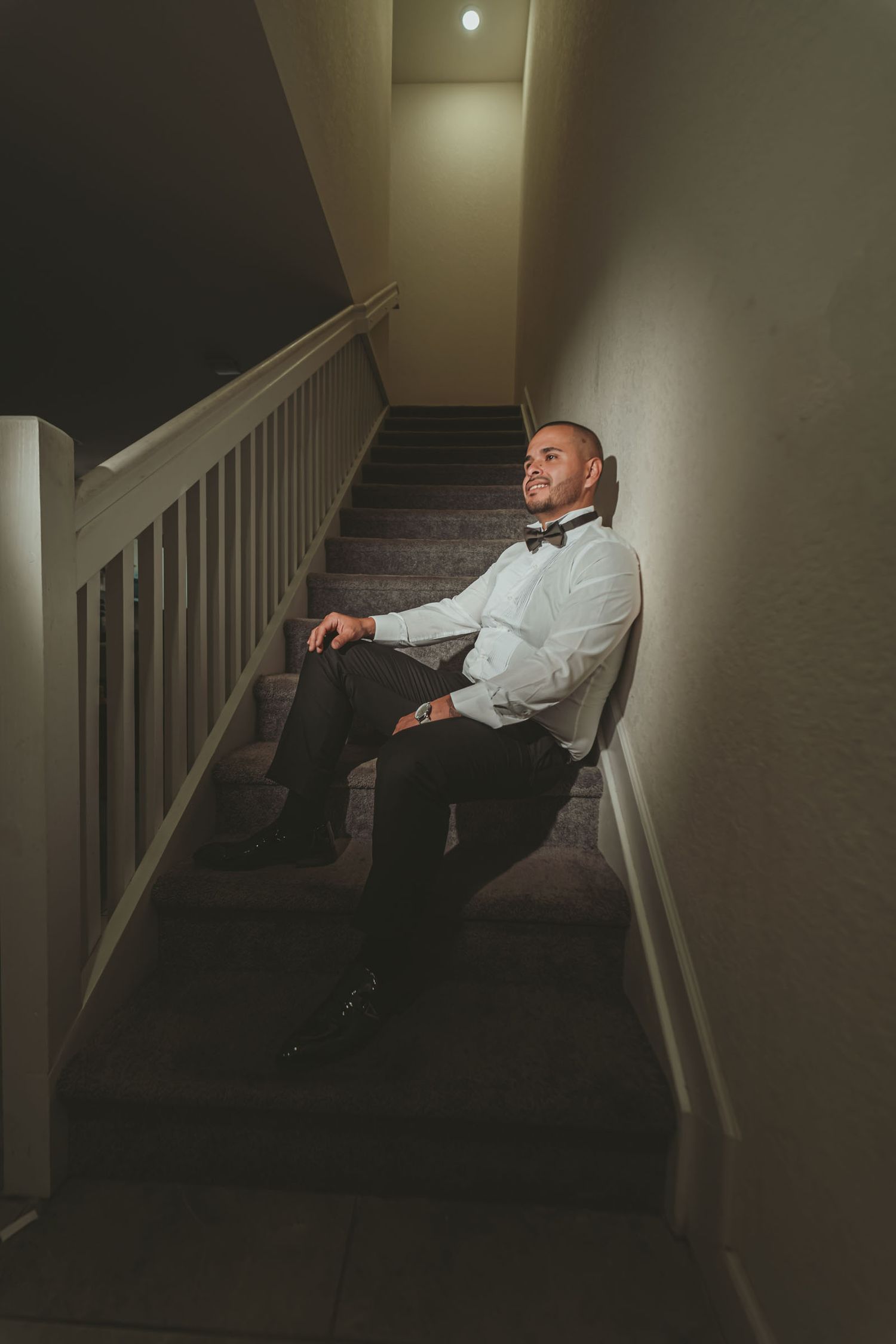 Novio con camisa y pajarita sentado relajado en escalera con luz tenue; preparación boda.