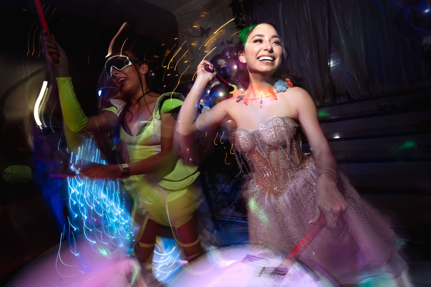 Quinceañera (vestido corsé) y animadora (traje neón) bailan con glow sticks en fiesta 'hora loca'; luces, movimiento.