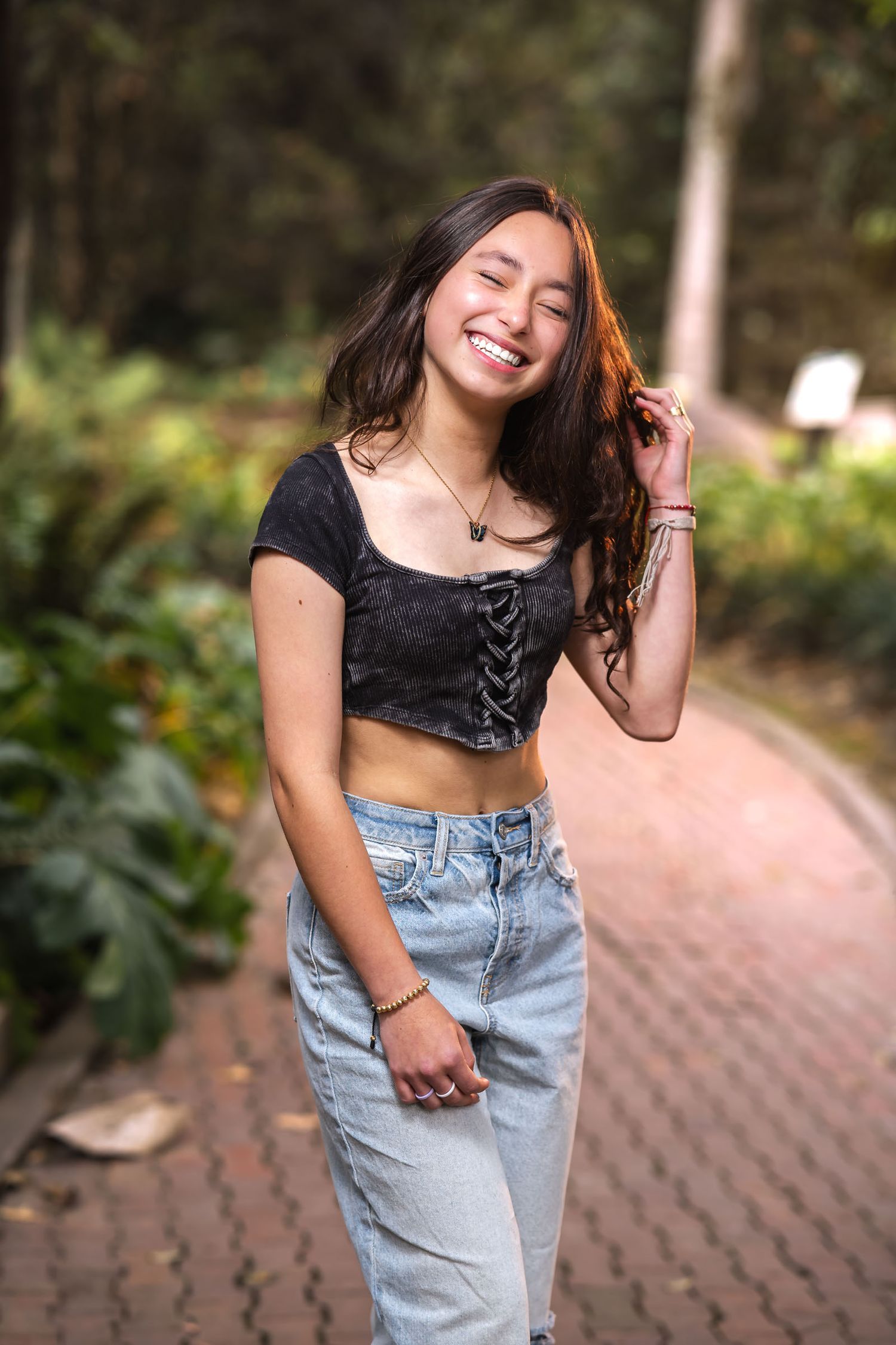 Joven sonriente con top negro corsé y jeans camina por sendero adoquinado en parque; sesión fotos.