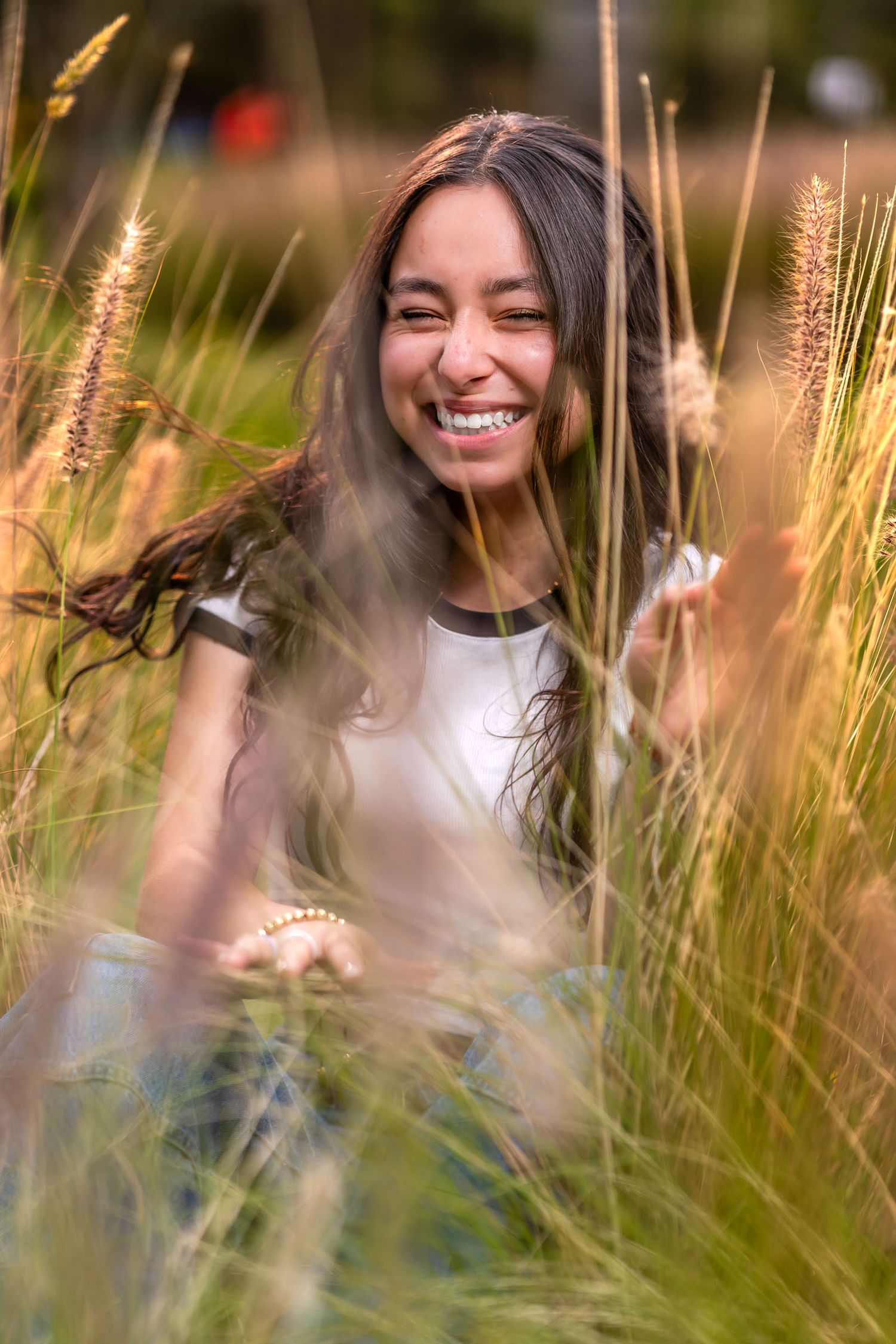 Joven ríe alegremente entre pasto alto dorado; foto espontánea vista a través de hierbas. Sesión prequince.