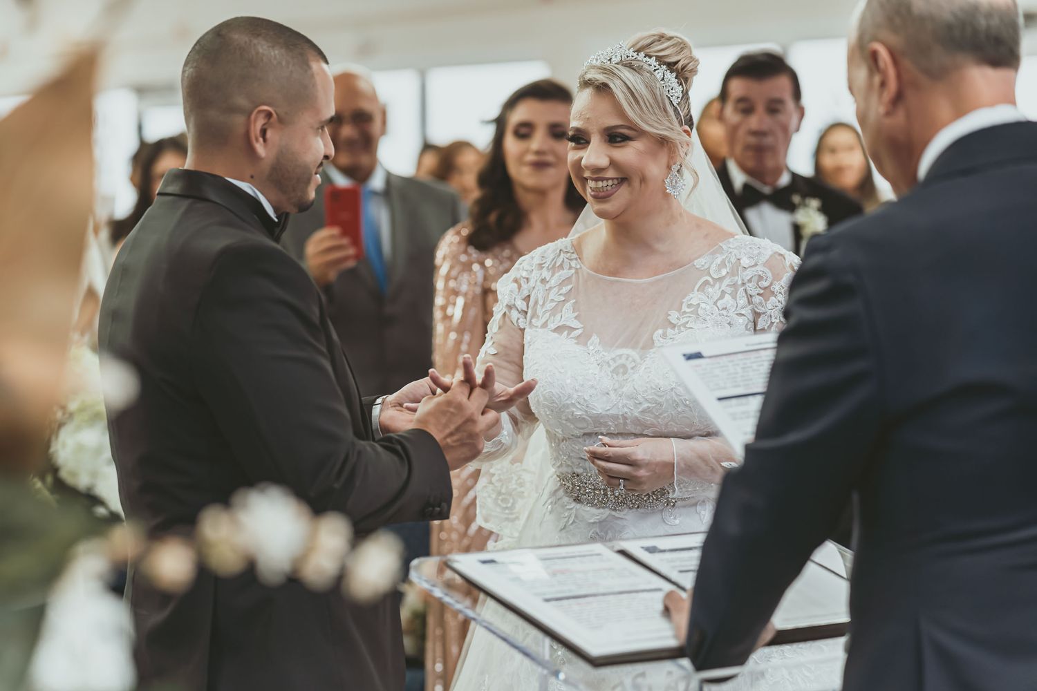 Novio coloca anillo en dedo de novia sonriente durante ceremonia boda; oficiante y documentos presentes.