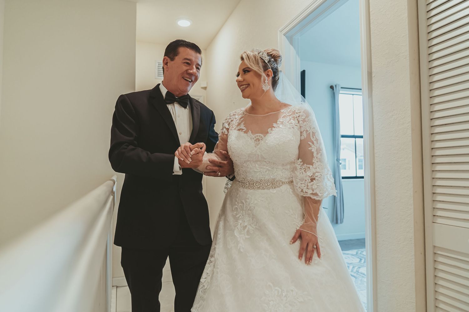 Novia con vestido blanco sonríe a su padre (en esmoquin) tomados de la mano en pasillo; momento emotivo boda.