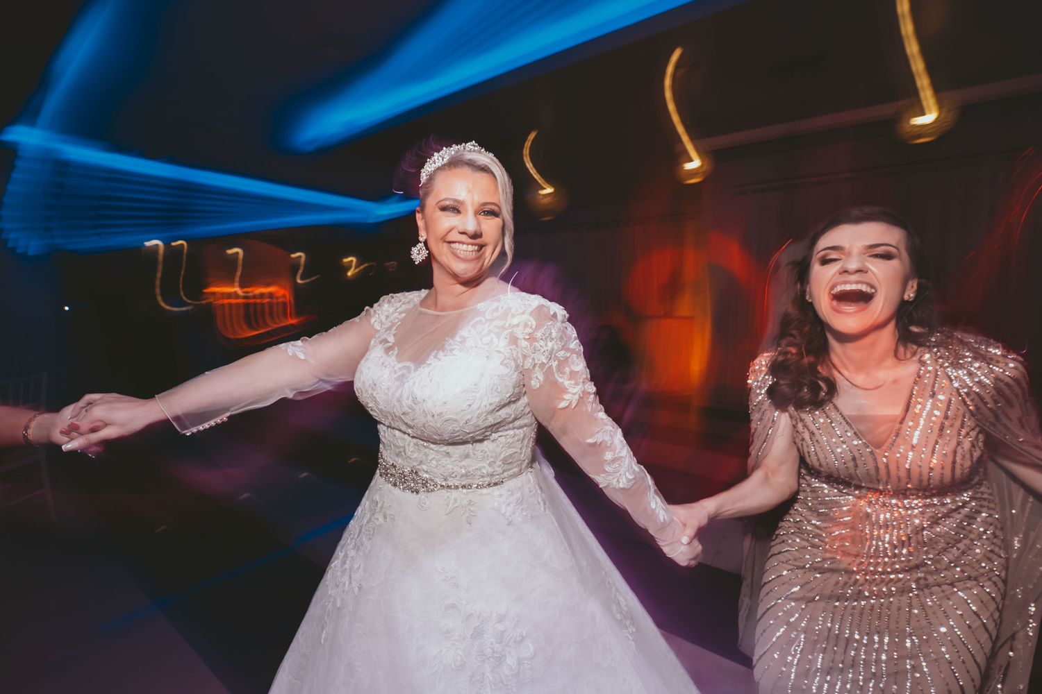 Novia sonriente con tiara baila tomada de mano con invitada; luces de fiesta coloridas y movimiento.