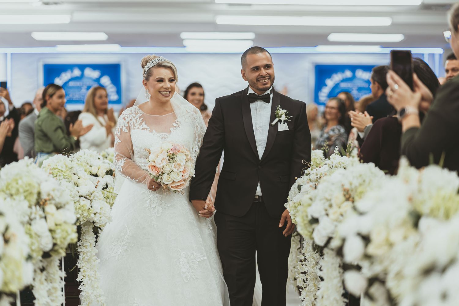 Novios sonrientes caminan tomados de la mano en salida nupcial por pasillo de iglesia; ella con tiara y ramo, él esmoquin. Boda idmji