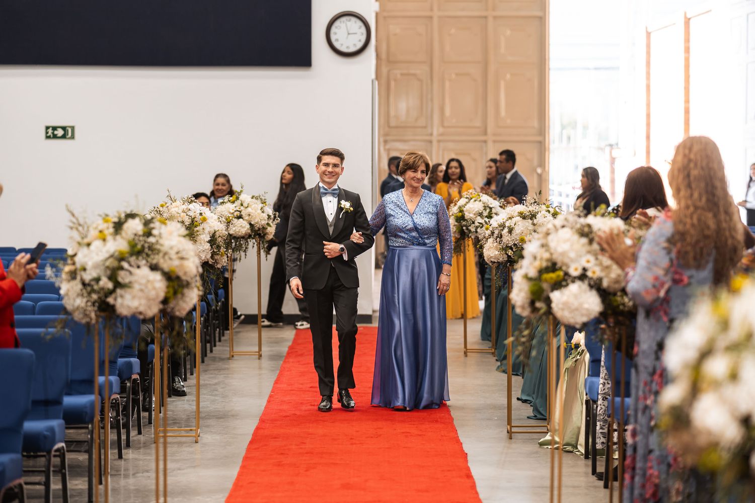 Novio con esmoquin camina del brazo con su madre (vestido azul) por pasillo con alfombra roja en ceremonia de boda.