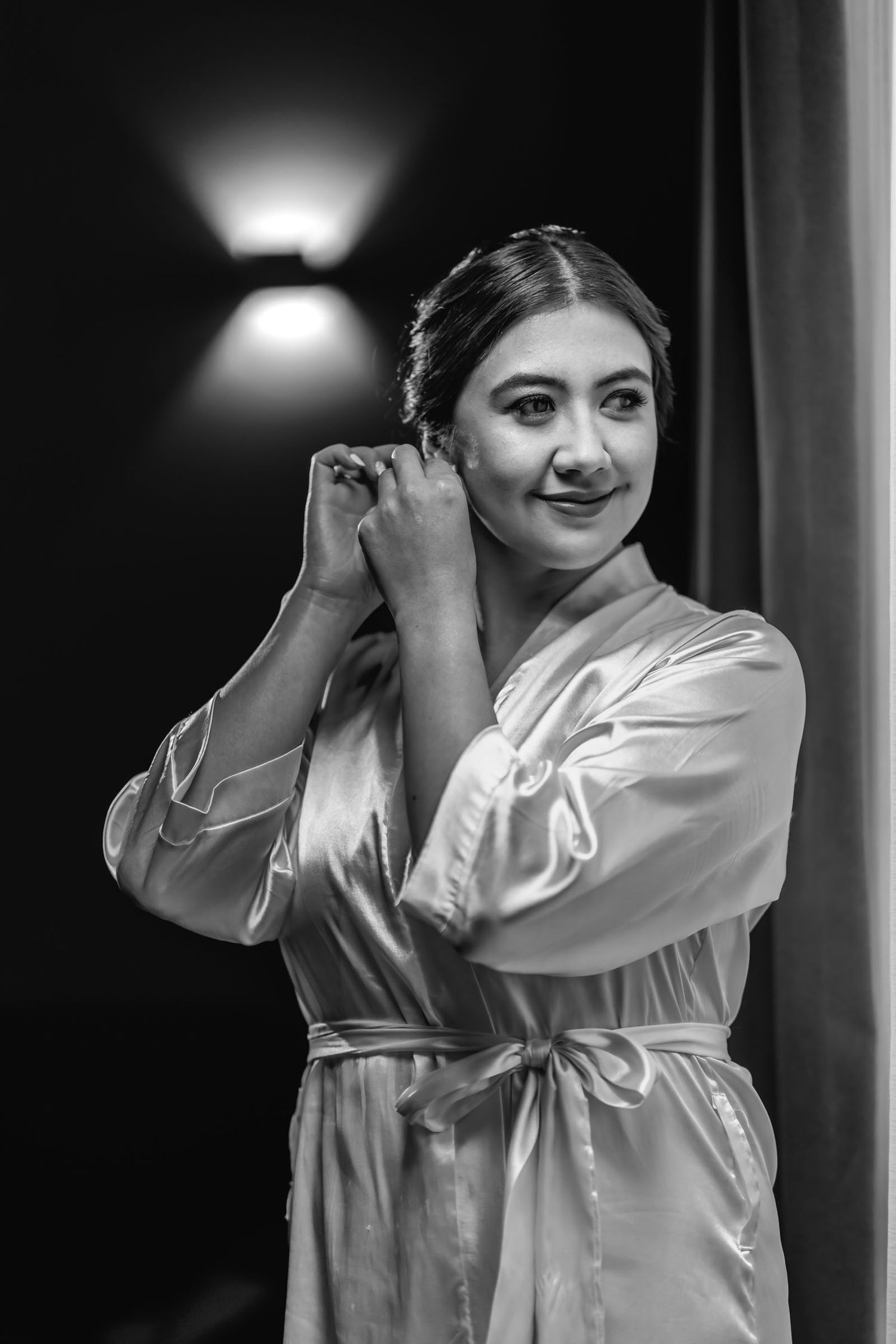 B&W: Novia con bata de satín se ajusta el arete sonriendo; preparación de boda, luz de pared al fondo.
