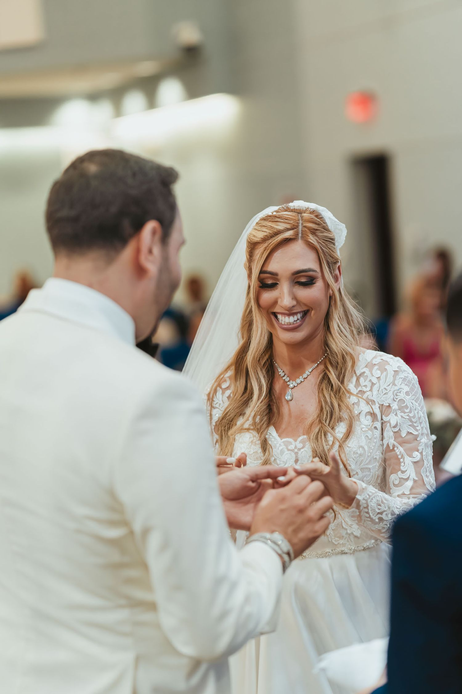 Novia sonríe feliz mientras novio (traje blanco) le coloca el anillo; ceremonia boda, invitados fondo.