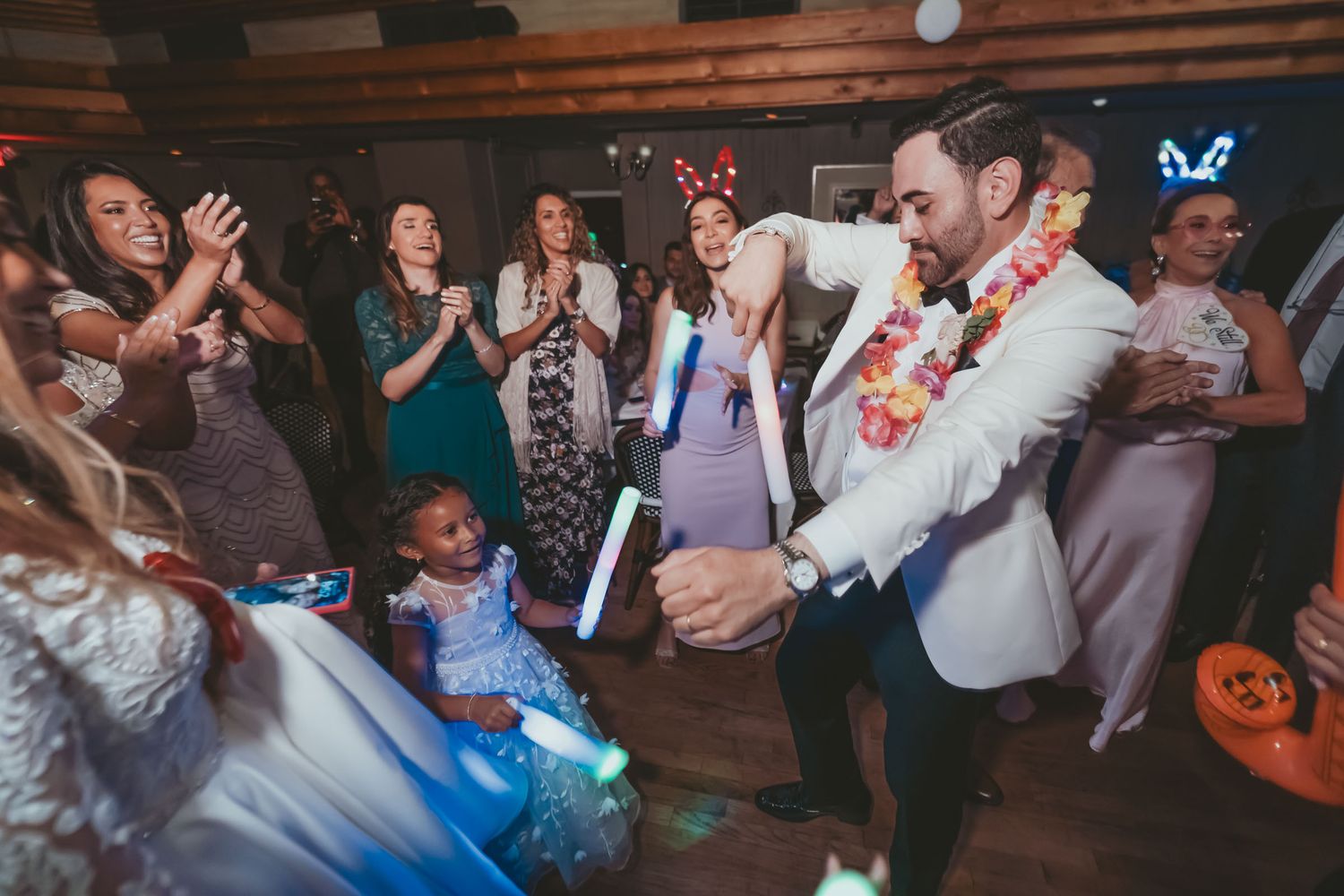 Novio con collar de flores baila animadamente con glow sticks en fiesta boda; invitados aplauden. Hora loca.
