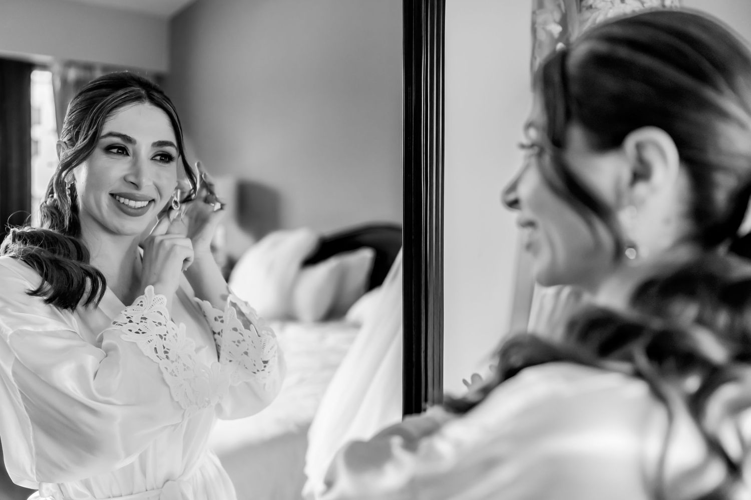 Foto B&W: Novia sonriente con bata blanca se ajusta el arete mirándose en espejo; preparación boda.