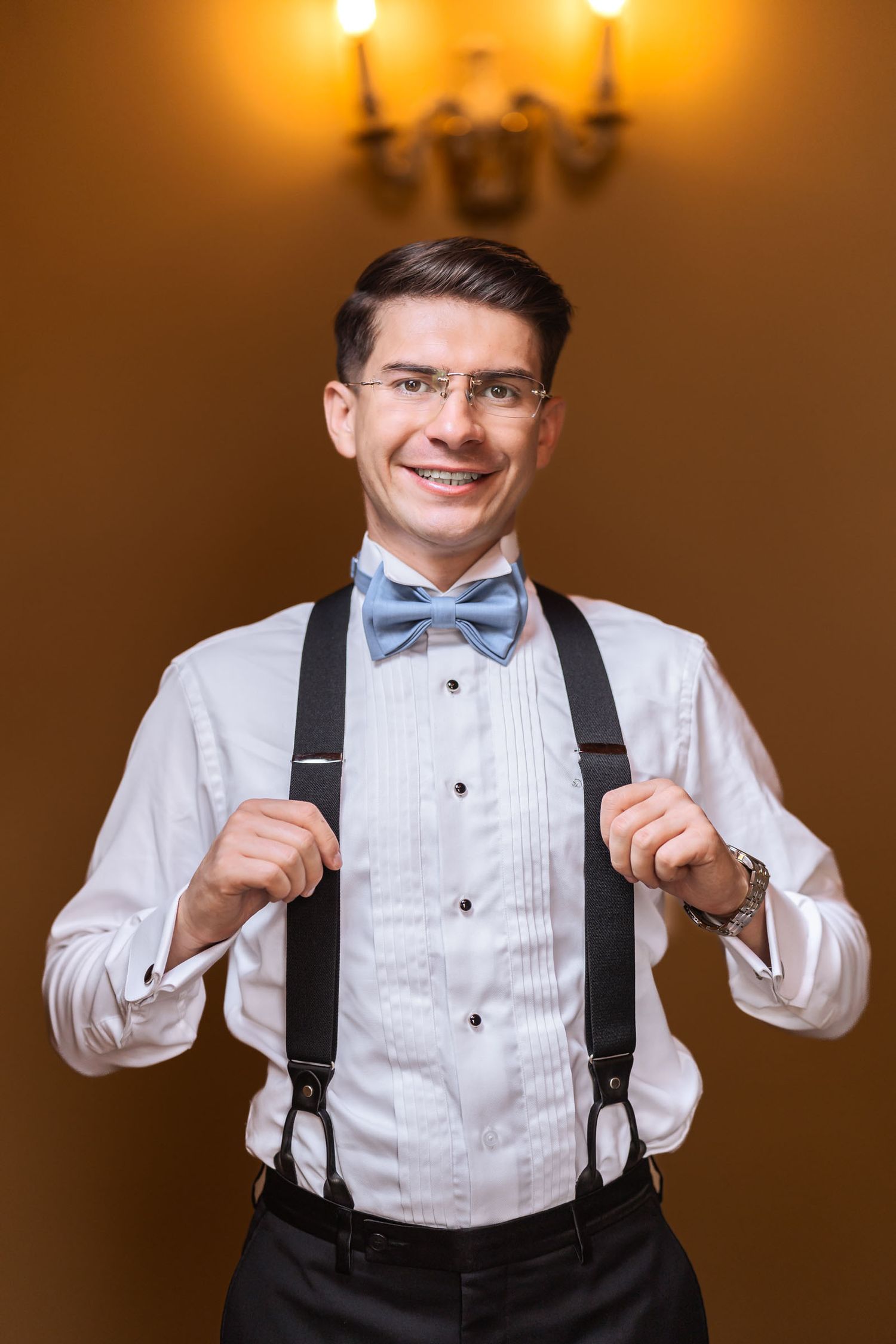 Novio sonriente con camisa blanca, pajarita azul y tirantes, posa sujetando los tirantes; preparación de boda.