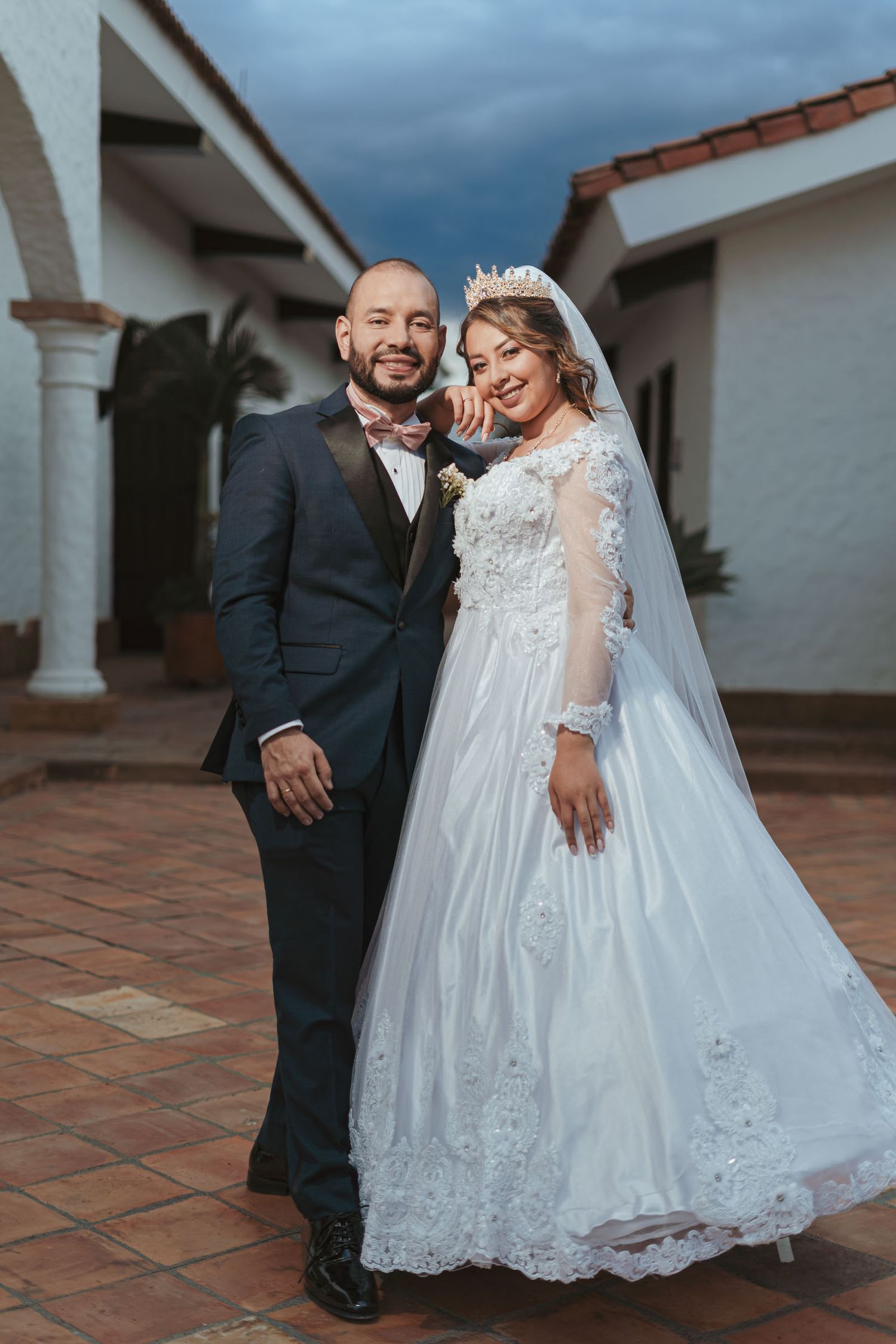 Recién casados sonrientes en Sáchica, Colombia. Novia con vestido blanco de manga larga y corona, novio con traje azul. Celebración alegre