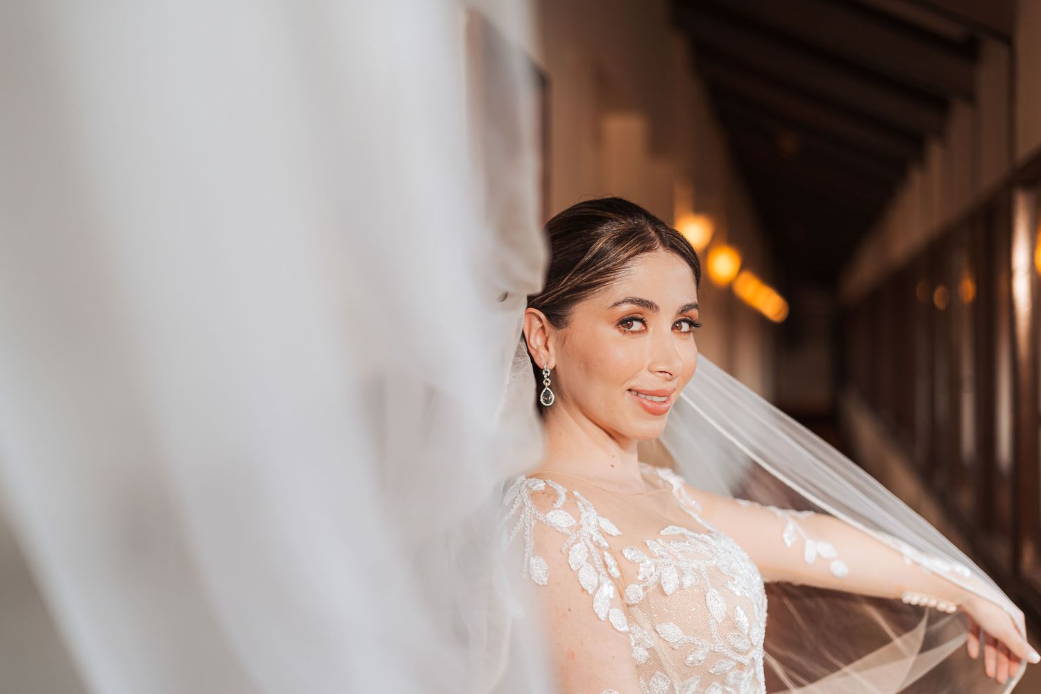 Novia sonriente en pasillo, velo largo flota a su alrededor; vestido bordado manga larga. Retrato boda.